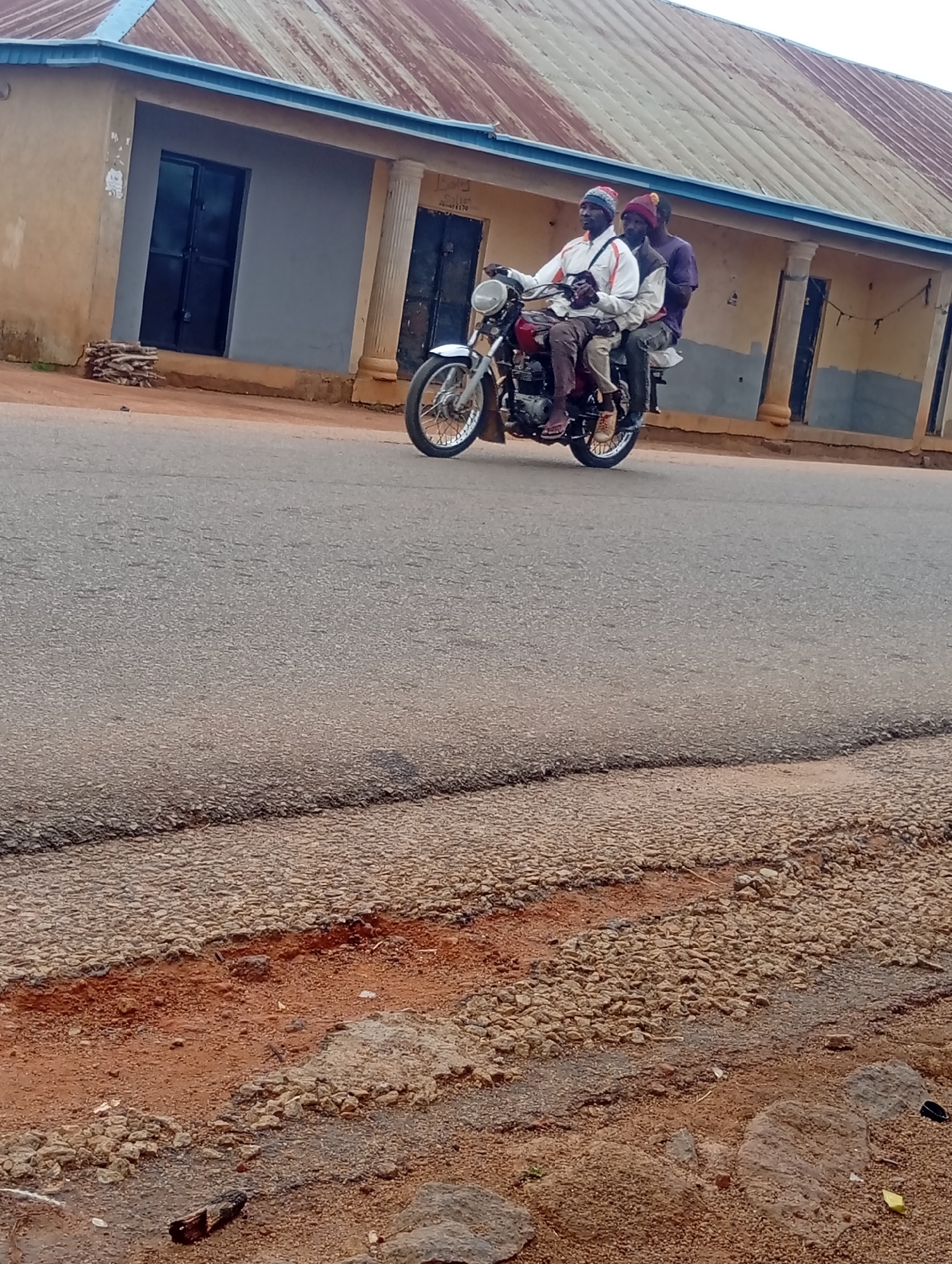 Three people ride a motorcycle on a paved road in Du, Nigeria. The road is cracked and damaged near the edge. Buildings with blue trim are along the roadside. The riders wear hats and casual clothing. The motorcycle is a red and silver model.