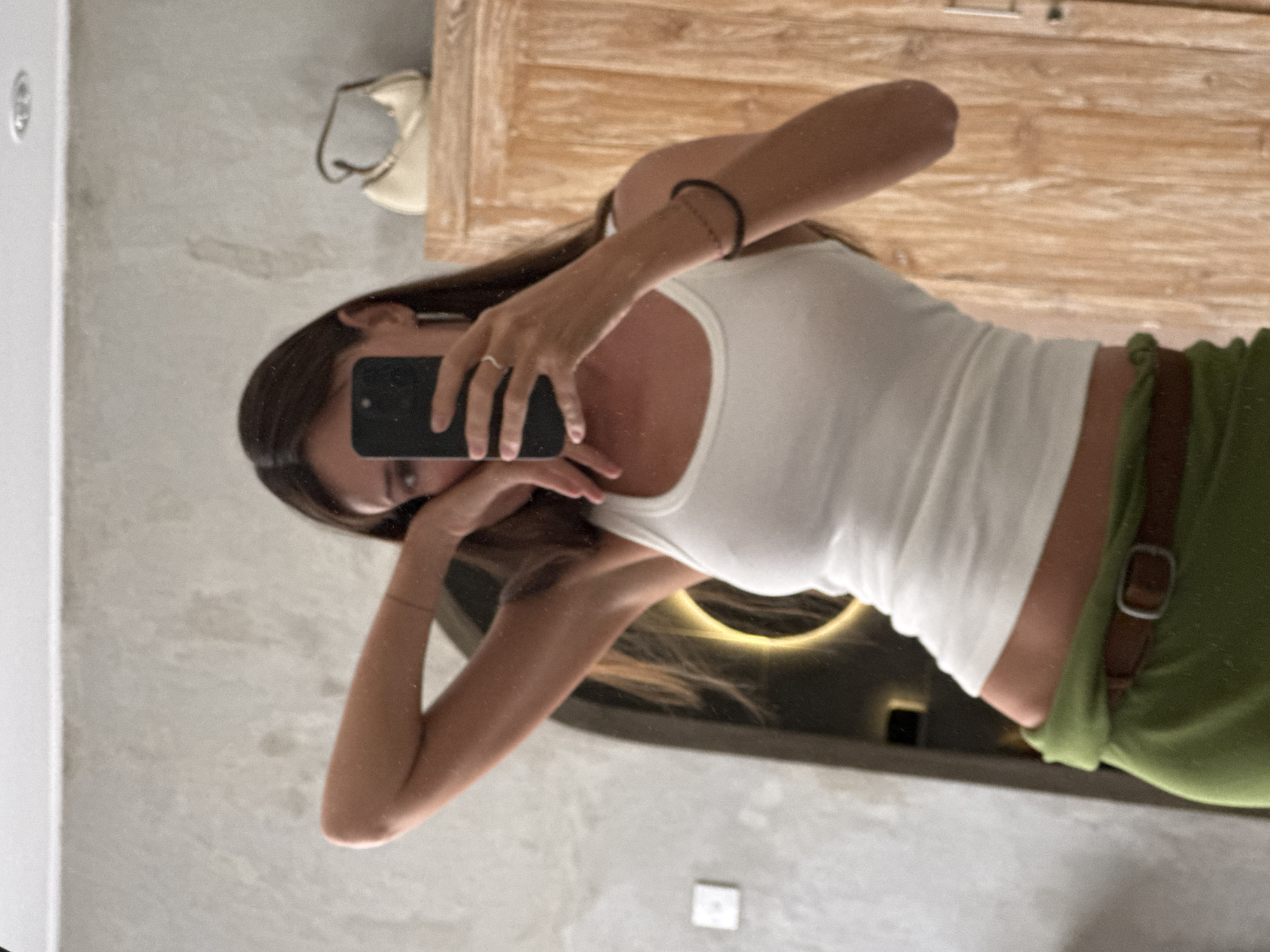 A woman is taking a mirror selfie in what appears to be a bathroom or dressing area. She is wearing a white tank top and green pants with a brown belt. She is holding a black smartphone in her right hand, angled to capture her reflection. Her left arm is bent and raised, with her hand resting near her face. The background shows a textured wall, a wooden cabinet with a light-colored purse on top, and a light fixture in the ceiling. There is also an arched doorway with a circular light source reflected within it. The overall impression is that of a private, domestic setting. The location is specified as North Kuta, Indonesia, suggesting a potential vacation or residence.