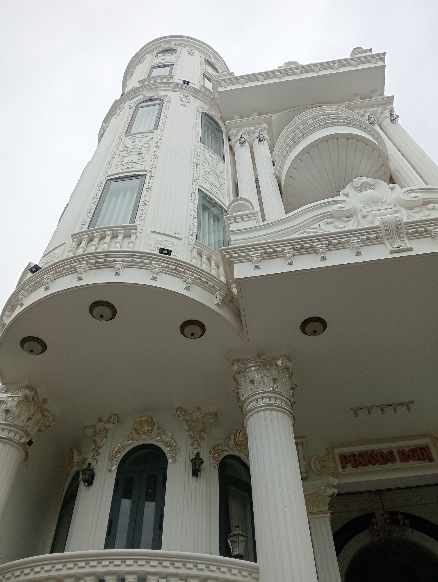 The image depicts the exterior of an ornate, white building in an unknown city. The architecture is characterized by classical elements, including decorative columns, balustrades, and embellished facades. A cylindrical tower with arched windows dominates one side, while the main entrance is marked by grand columns with intricate capitals. Lighting fixtures are visible on the underside of balconies and overhangs. The weather appears overcast, and the sky is a uniform gray. A sign is visible near the entrance, though the specific wording is partially obscured. The overall impression is one of ostentatious wealth and elaborate architectural design.