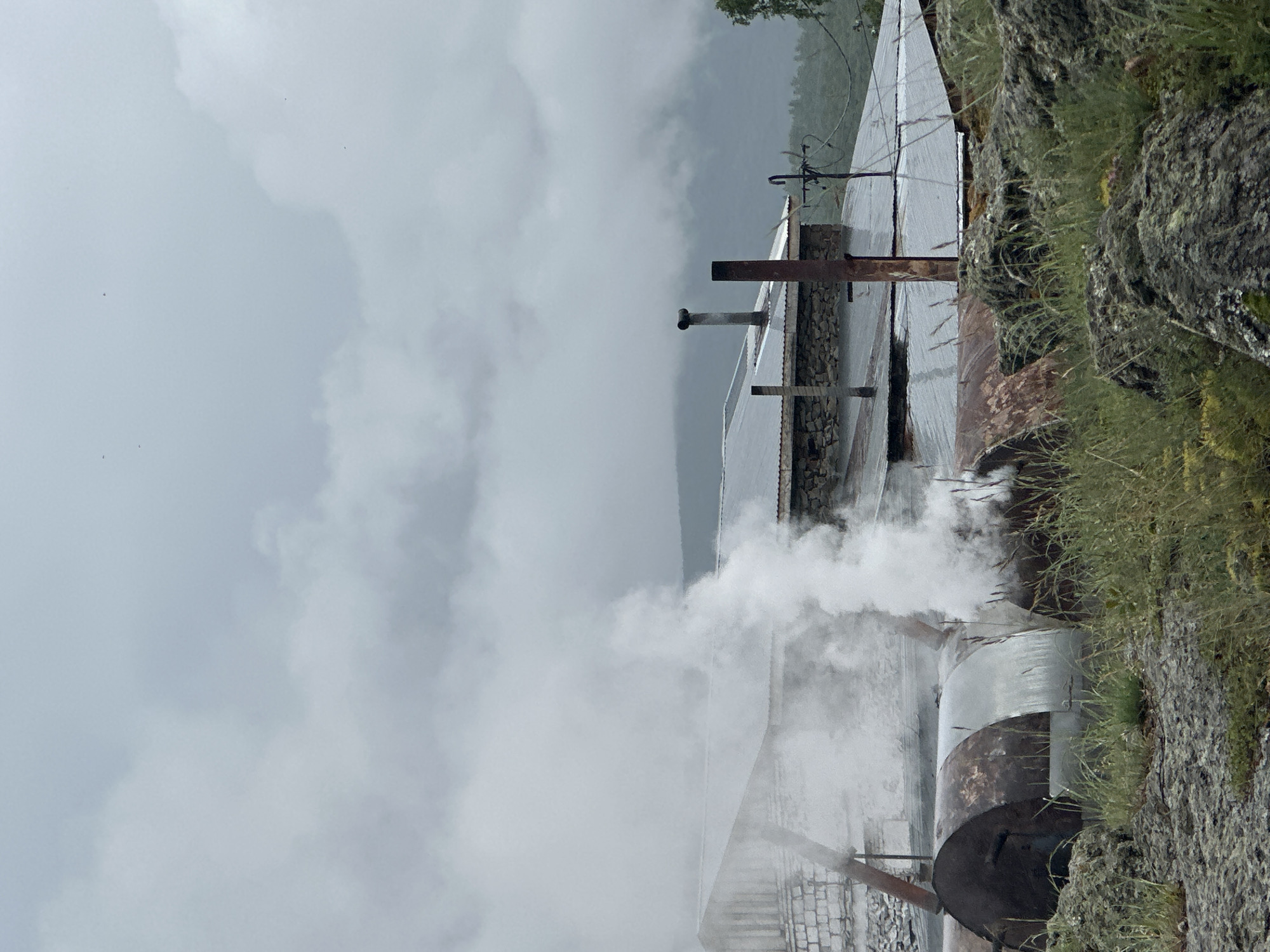 An outdoor scene shows industrial structures with smoke billowing upwards under a cloudy sky. In the foreground, large, rusted metal barrels are visible, with steam emerging from at least one of them, suggesting an active process. The barrels are situated on rocky terrain with dry grass. Behind the barrels, several buildings with corrugated metal roofs are present. One building appears to be constructed from white bricks and stone. Chimneys are visible on the roofs of some buildings, and electrical wires and a utility pole are also part of the scene. In the background, a misty, mountainous landscape is present, fading into the overcast sky. The overall impression is one of an industrial operation in a rugged, possibly remote, environment.