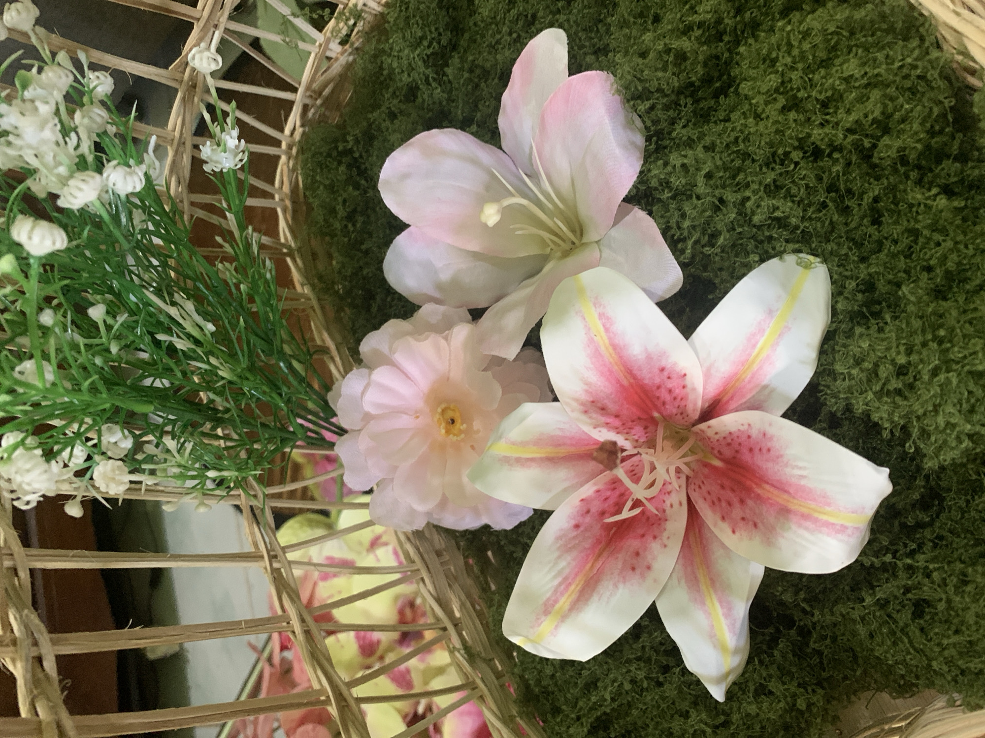 The image displays a close-up view of a decorative arrangement inside a woven basket. The basket is made of light-colored reeds and contains a layer of green, moss-like material. On top of the moss are three artificial flowers. The largest flower in the foreground is a lily with white petals heavily speckled with pink and streaks of yellow. To its right is another large, light pink lily with white accents. Behind these, a smaller, fluffy pink flower with a yellow center is partially visible. Behind the flowers, a cluster of artificial greenery and tiny white flowers can be seen, also within the basket. The background is blurred, showing hints of more woven material and a dark, indistinct surface. The scene appears to be an indoor decorative display, potentially for an event or as part of home decor. There are no people or animals present. The image does not provide any specific clues about its location in Kuala Pembuang, Indonesia.