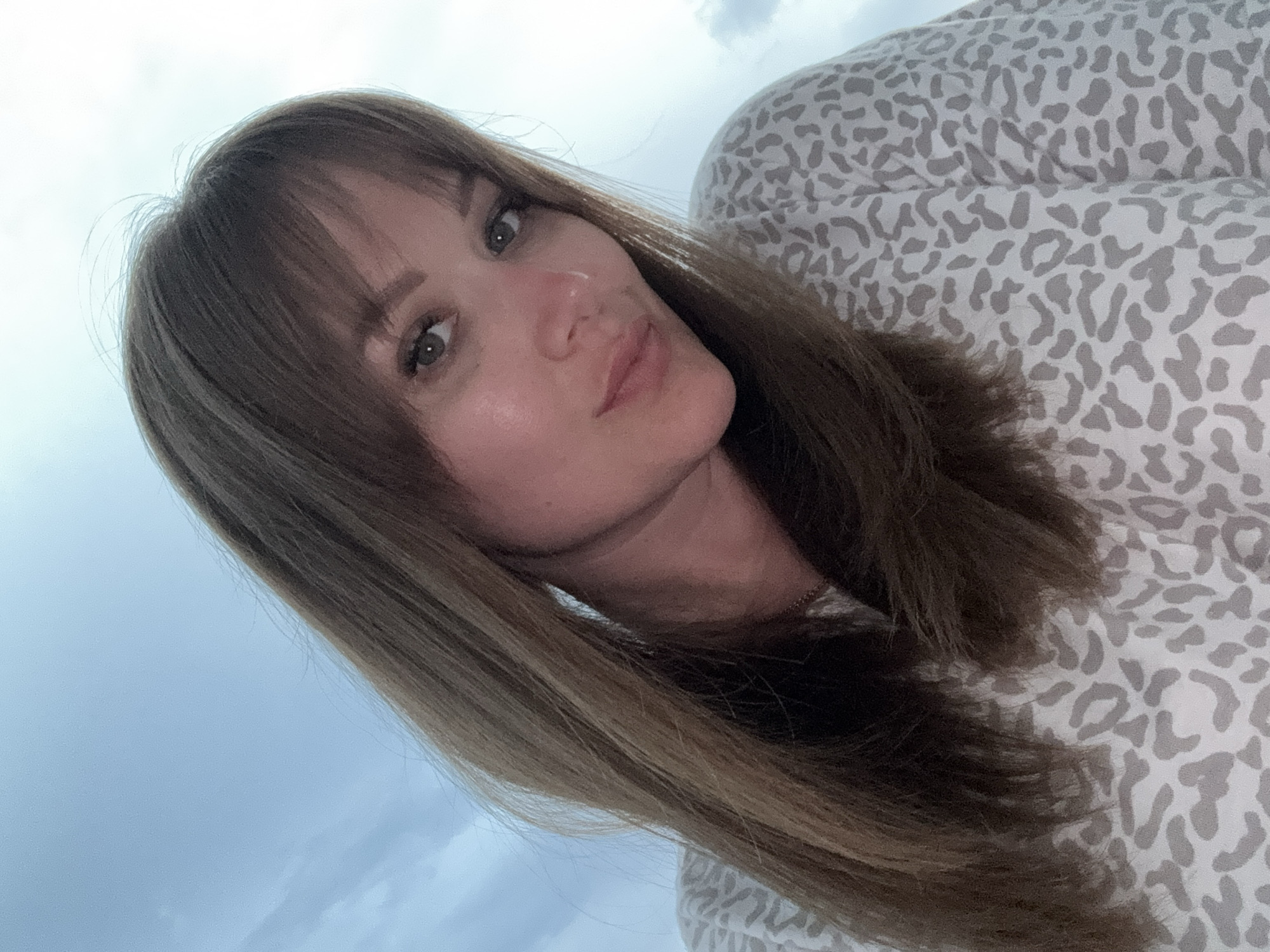 A woman with medium-length brown hair and bangs is centered in the frame. She is wearing a light-colored shirt with a gray animal print. The background consists of the sky, which is a light blue-gray color. There is no indication of this being in the city of Yevpatoriya, Ukraine.