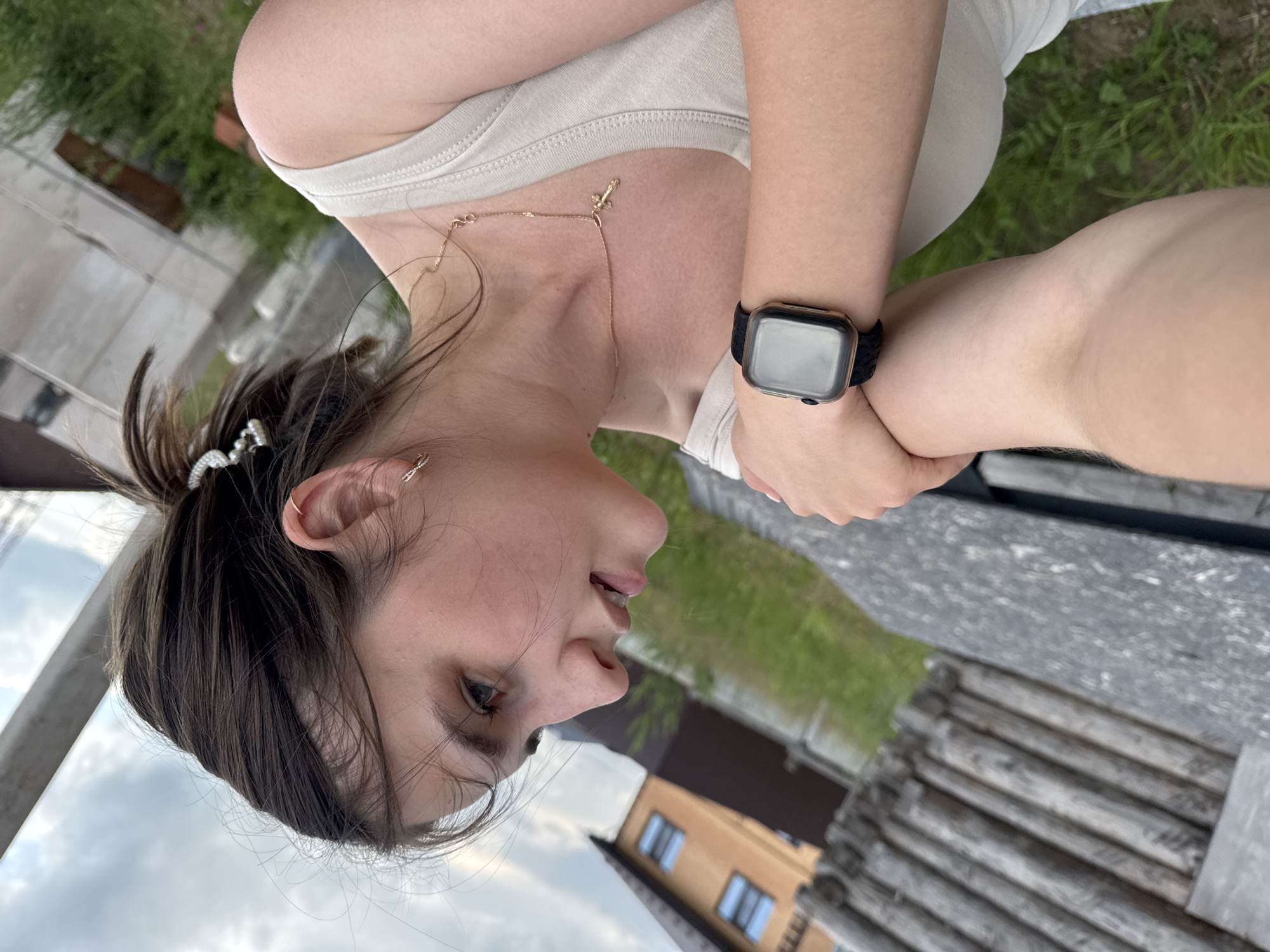 The image shows a young woman with brown hair, wearing a beige tank top and a watch on her left wrist. She is looking upwards and to the right. In the background, there is a wooden structure and a building that appears to be a house. The location is identified as the city of Chaikovsky, Russia, and the scene is set outdoors with green grass and a cloudy sky. The woman is also wearing a necklace with a cross pendant and small earrings. The composition suggests a casual, outdoor setting.