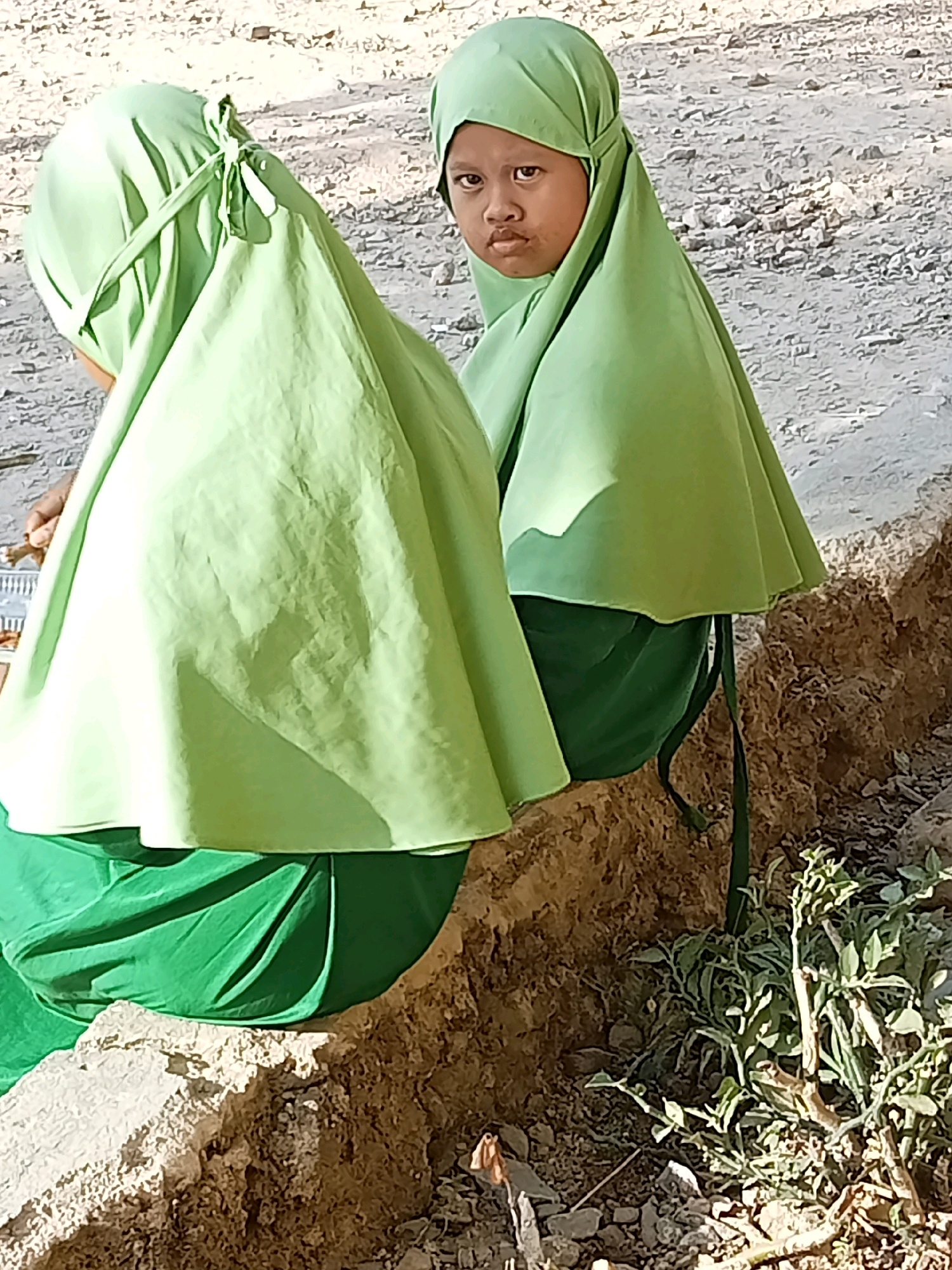In Lebo Jaya, Indonesia, two young individuals are seated outdoors on a low stone wall. One individual, facing the camera, wears a light green hijab and a dark green garment. Their gaze is directed towards the viewer with a neutral expression. The other individual, partially visible from the back, also wears a light green hijab and a matching garment. The background consists of dry, dusty ground with scattered pebbles and sparse vegetation. The lighting suggests it is daytime, with shadows indicating the sun's position.