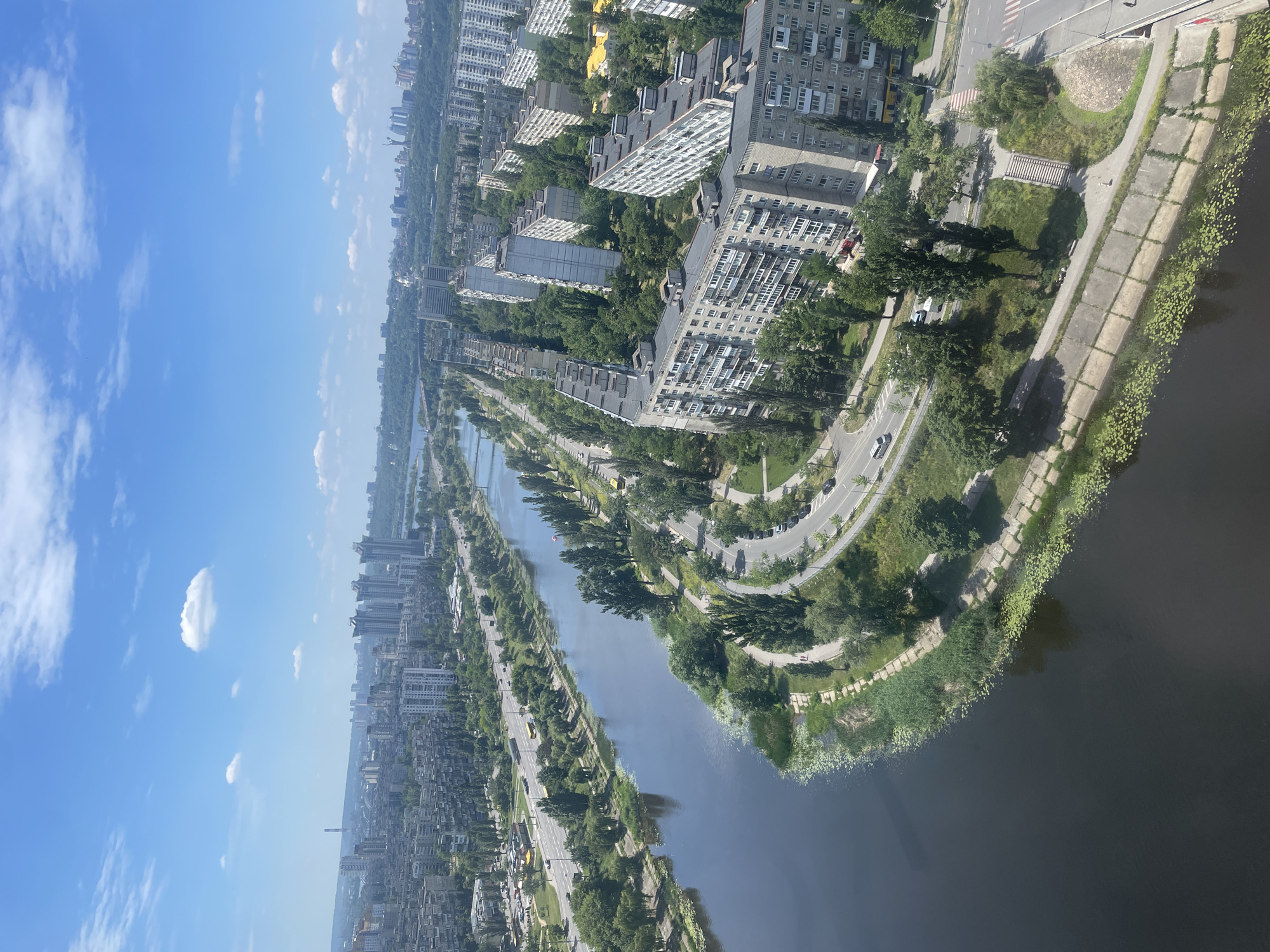 An aerial view captures a sunny day in Kyiv, Ukraine, showcasing a wide river curving through a dense urban landscape. The river, likely the Dnieper, dominates the foreground with its dark, still water, bordered by a concrete embankment and a strip of lush green vegetation. Beyond the river, a multi-lane road runs parallel to its bank, with several cars and a yellow bus visible. Across the road, a series of large, uniform apartment buildings, typical of Soviet-era architecture, stretch along the cityscape. These buildings are interspersed with numerous tall, mature trees, creating a green canopy over the residential areas. In the distance, more buildings and a few bridges spanning the river are visible, fading into a hazy horizon under a bright blue sky dotted with scattered white clouds. The scene depicts a vibrant, residential part of Kyiv, characterized by its river, expansive roadways, and extensive greenery amidst high-rise housing.