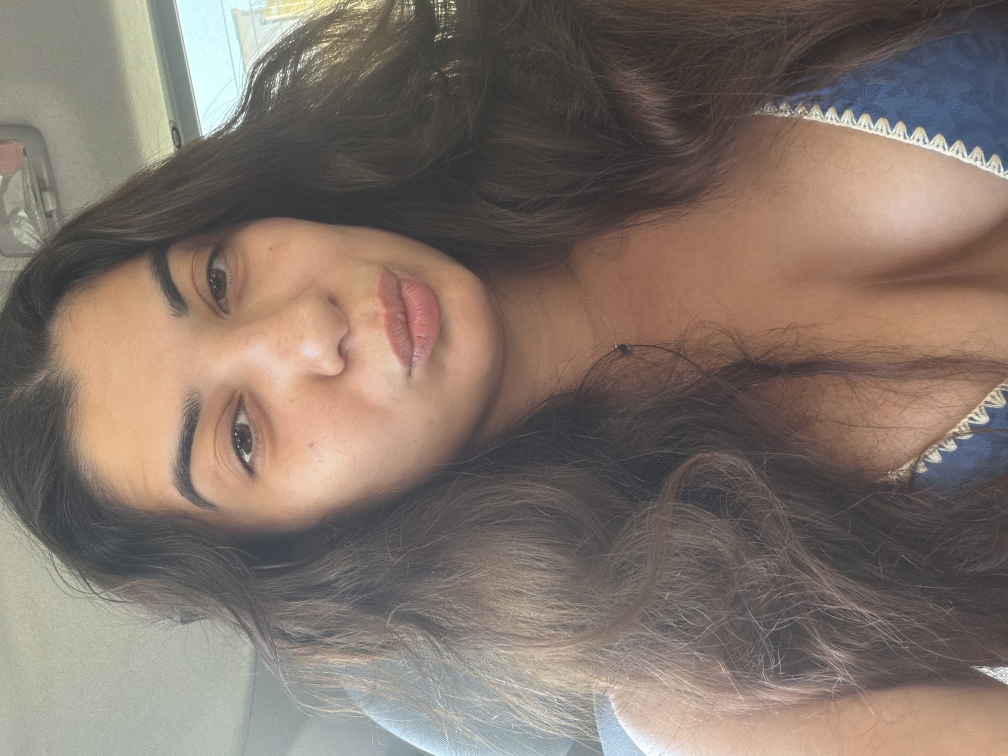 A woman with dark, wavy hair is shown in a close-up, selfie-style photograph, likely taken inside a vehicle. She is looking directly at the camera with her lips puckered. She is wearing a blue top with white trim. The background is blurred but appears to be the interior of a car, with a rearview mirror visible at the top. The lighting suggests it is daytime.
