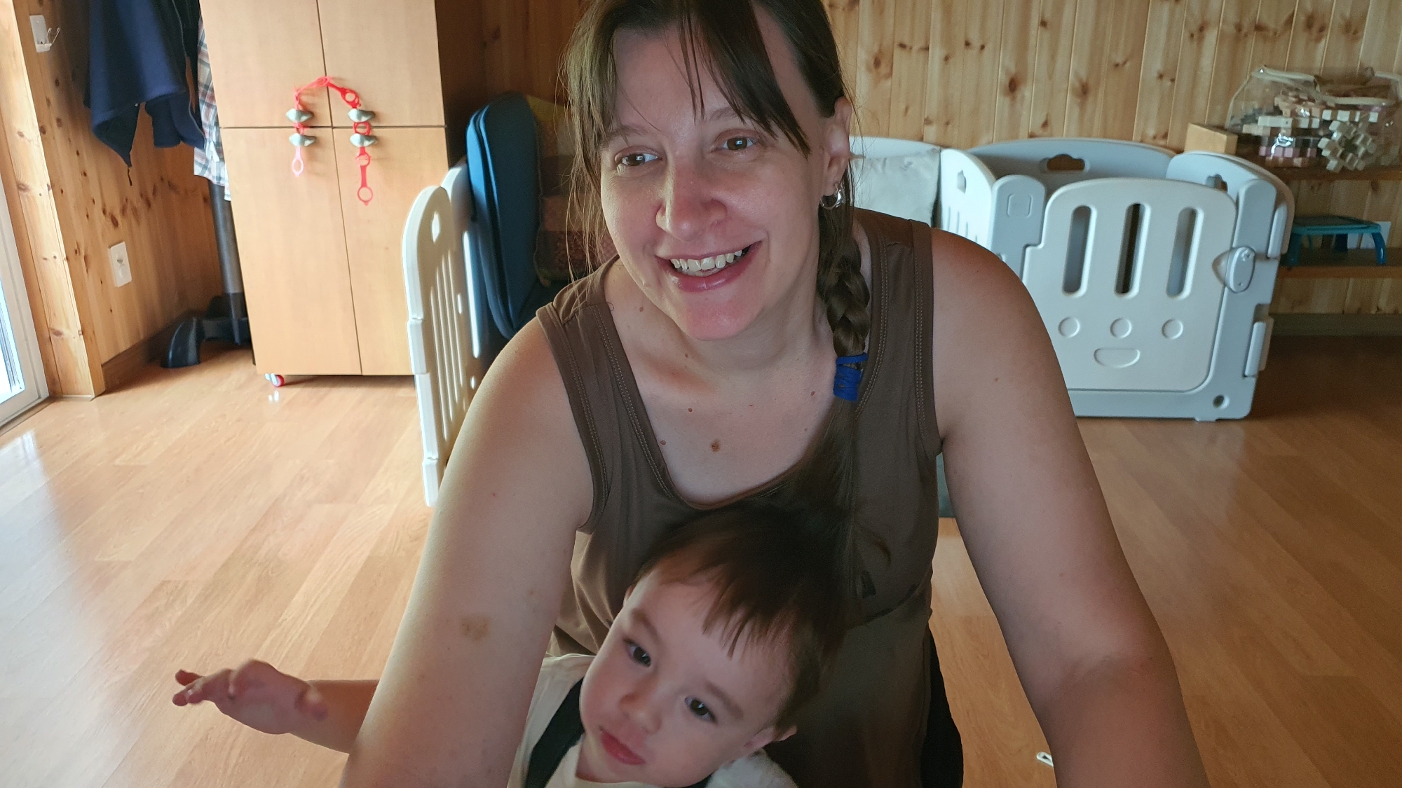 A woman with brown hair and a brown tank top smiles at the camera while holding a baby. The baby is wearing a white shirt with black suspenders and has brown hair. The woman's hair is pulled back in a braid with a blue tie. They are in a room with wooden walls and floor. In the background, there is a tall wooden cabinet with red decorations on it, a blue laundry basket, and a white plastic playpen. There are also shelves with toys in the background. The lighting in the room is bright, suggesting it is daytime.