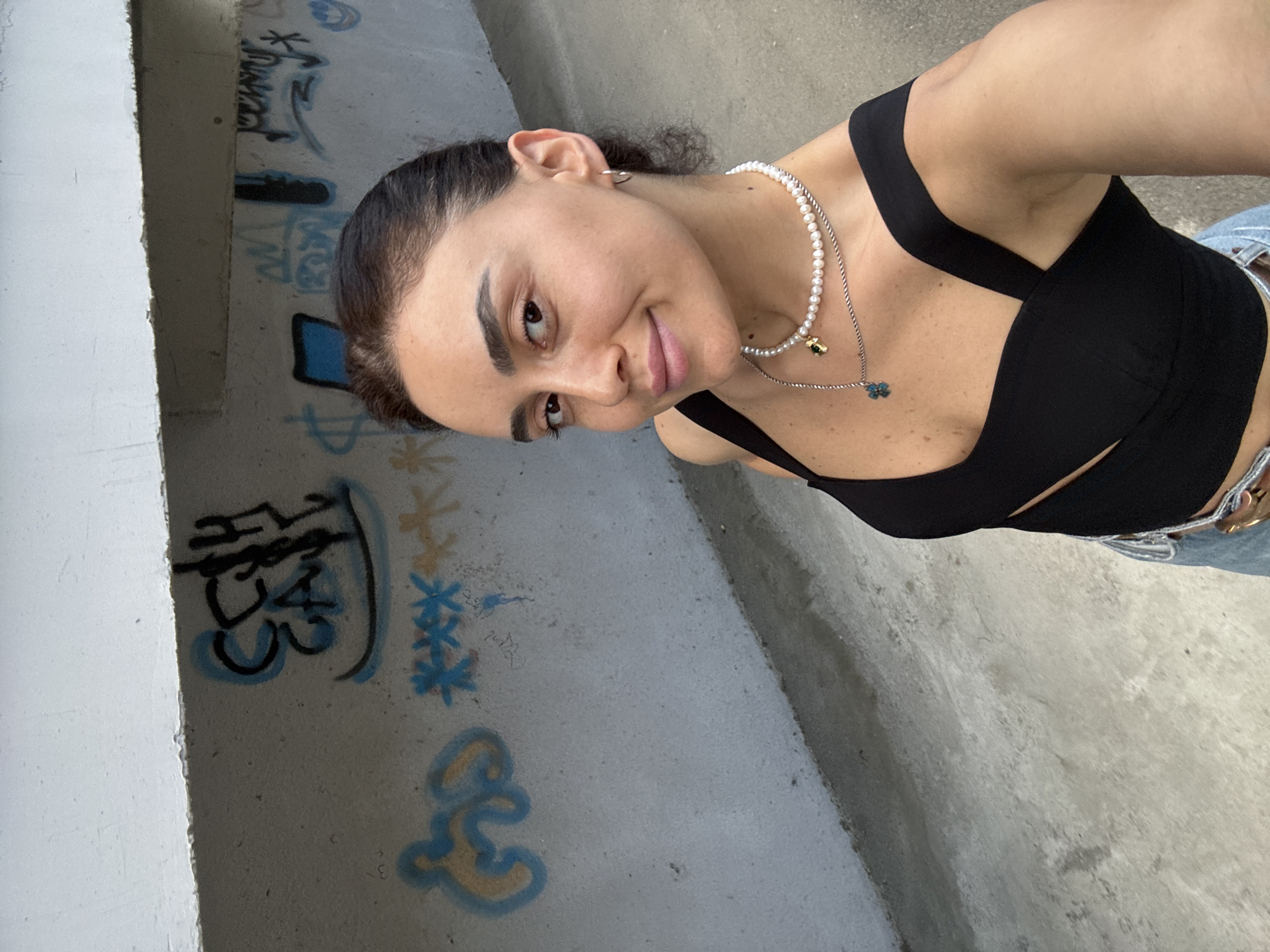 The image shows a young woman with dark hair and brown eyes, wearing a black crop top and jeans. She is looking at the camera and smiling slightly. She is also wearing a pearl necklace with a gold charm. The background is a concrete wall with graffiti on it. The graffiti includes various symbols and writing in different colors, including blue, yellow, and black. One of the graffiti appears to be Chinese characters. The scene is lit by natural light, suggesting it is daytime. The overall mood of the image is casual and confident. The woman's attire and the graffiti in the background suggest an urban setting. The image is a selfie, taken from a slightly high angle. The presence of graffiti and concrete walls hints at an outdoor urban environment, possibly a skate park or an underpass. The sunlight casts soft shadows, indicating it is likely late afternoon or early morning. The woman's relaxed posture and smile convey a sense of ease and self-assurance.