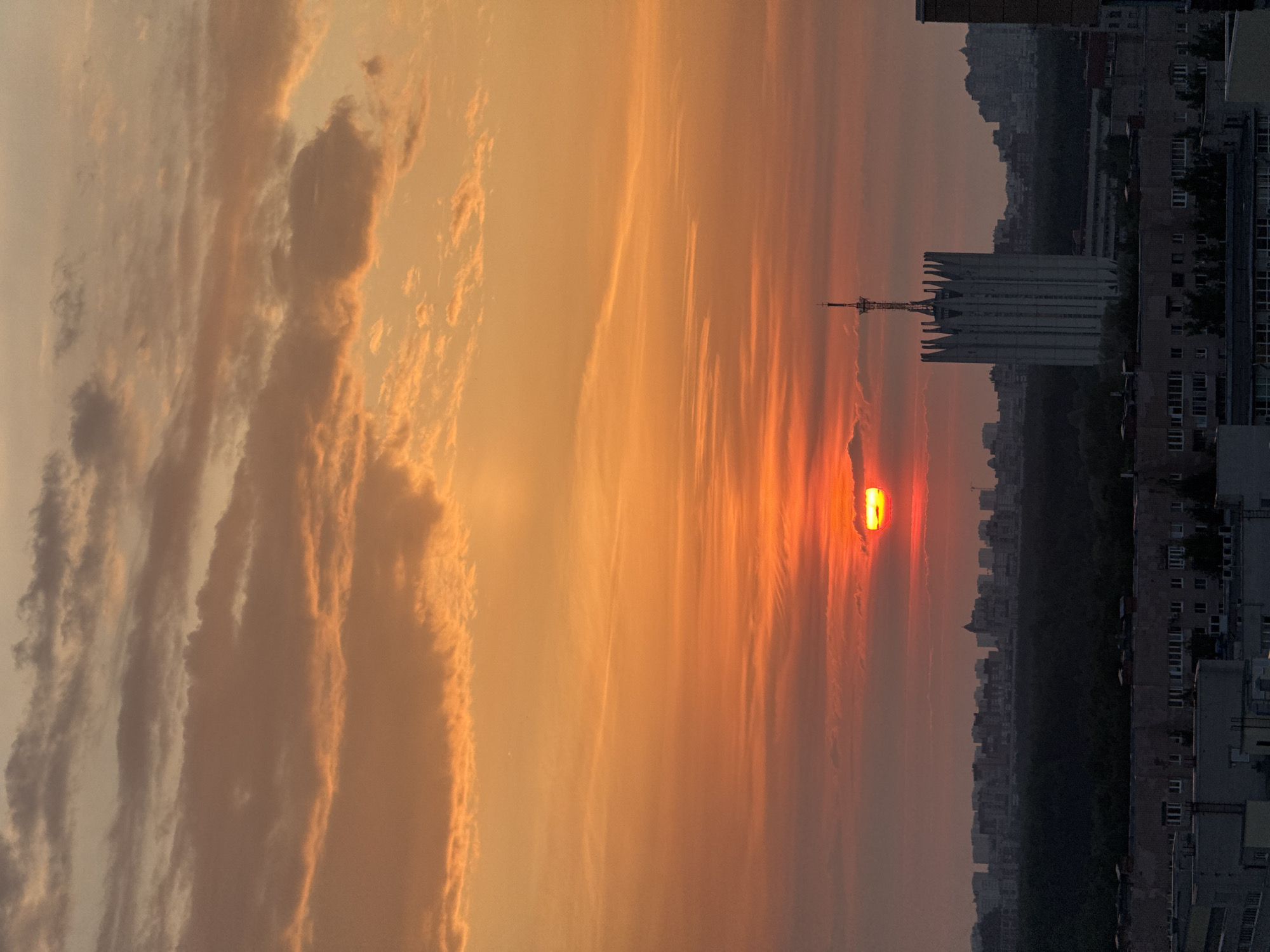 The image shows a sunset over a city, possibly Sinyavino, Russia. The sun is a vibrant orange color and is partially obscured by thin clouds. The sky is filled with orange and yellow hues, fading into a more muted gray color near the top. Buildings are visible in the background, appearing as dark silhouettes against the bright sky. A tall structure with a broadcast tower is a prominent feature of the skyline. The image is oriented vertically, with the sky dominating the frame.