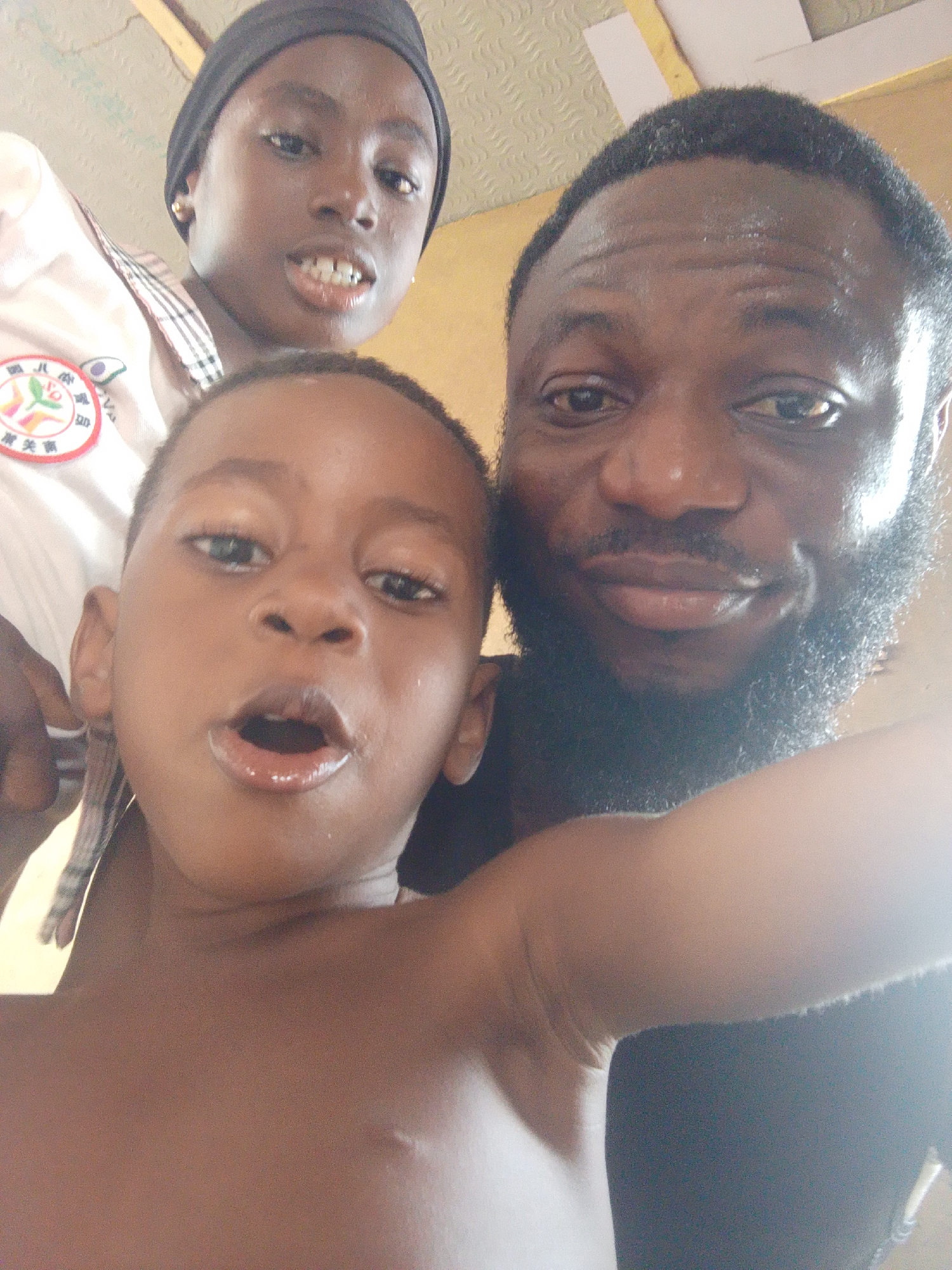 A close-up selfie taken indoors in Lokoja, Nigeria, features three individuals of African descent. On the right, an adult male with a dark, full beard and short dark hair gazes at the camera with a subtle smile. Behind and slightly above a young child, a younger person wears a dark head covering and a light-colored collared shirt with plaid accents. She also looks towards the camera, showing a slight smile with visible teeth. Her shirt bears a round, red and white emblem featuring colorful shapes (green, yellow, red) and Chinese characters, specifically "童星园" (Tóng Xīng Yuán, meaning "Children's Star Garden" or "Child Star Park") and "関商" (Guān Shāng). In the foreground, center-left, a young, bare-chested child with wide eyes has an open mouth, looking directly at the camera, conveying a sense of surprise or playful engagement. The background shows an indoor ceiling composed of light-colored panels with visible yellow structural elements. The overall bright lighting suggests it is daytime. The individuals appear to be posing casually for the photograph, capturing a moment of interaction or family connection.