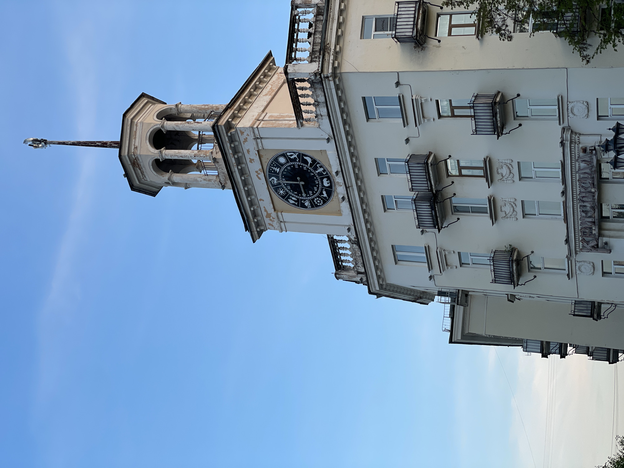 The image shows a tall building with a clock tower in Zheleznogorsk, Russia. The clock face features zodiac symbols instead of numbers. The building is white with multiple stories and small balconies. The sky is blue with some visible electrical wiring on the right.