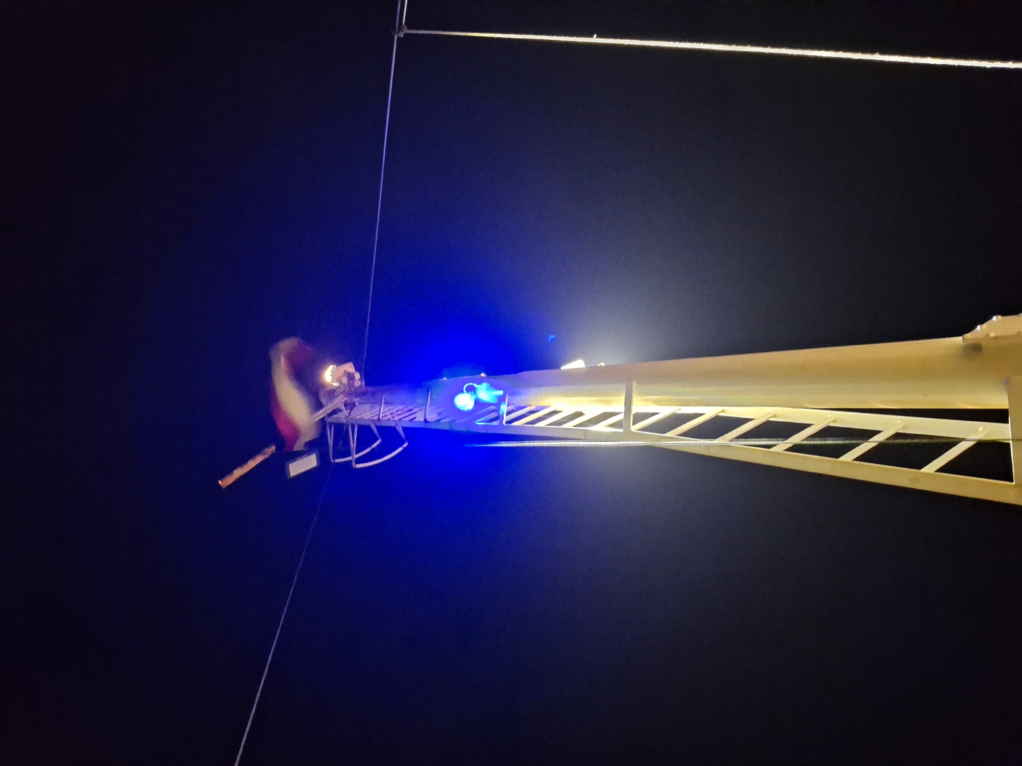 A low-angle shot depicts a fire truck ladder extended into a dark night sky. The ladder's end features a rescue platform with flashing blue lights. At the edge of the platform, a figure in a fire helmet and gear is visible. A thin cable stretches diagonally across the frame. The location is Unknown, Unknown.