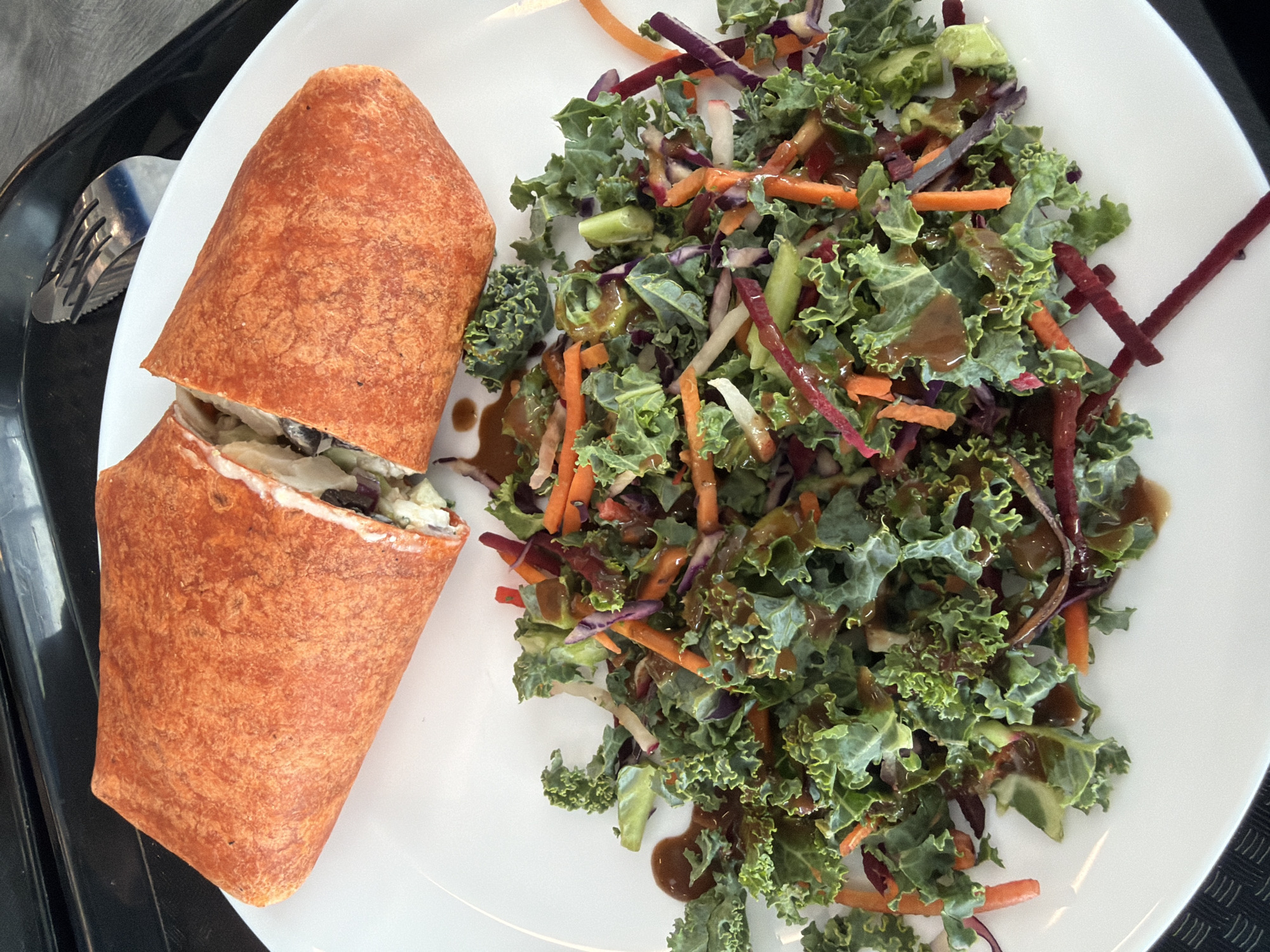 The image displays an overhead view of a meal served on a white plate, resting on a black tray, presumably in Mirabel, Canada. The primary subjects are a large, reddish-orange wrap and a side salad. The wrap is cut into two segments, revealing a filling of shredded light-colored vegetables, darker elements, and a creamy white dressing. The salad consists of dark green, curly kale leaves, julienned orange carrots, shredded purple cabbage, and thin white vegetable strips, all covered in a glossy, brown dressing. A silver fork with a textured handle is partially visible on the black tray in the upper right background. No people, actions, or interactions are visible.