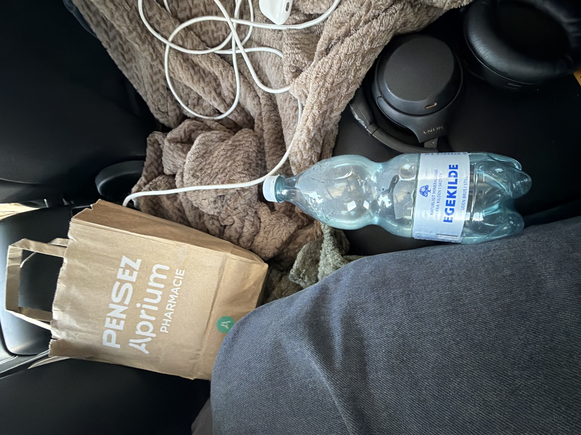 The image, taken inside a vehicle in Ödsmål, Sweden, shows a brown paper bag with the text "Pensez Aprium Pharmacie" printed in white. Next to the bag is a brown, fuzzy blanket. A clear plastic bottle of "EgeKilde" mineral water, labeled "UDEN BRUS / STILL," is positioned in front of the blanket. A pair of black Sony headphones rests on the blanket, and a white cable with an earbud is also visible. The lower left portion of the image displays a person's dark gray denim-clad leg. The scene appears to be a personal collection of items within a car.