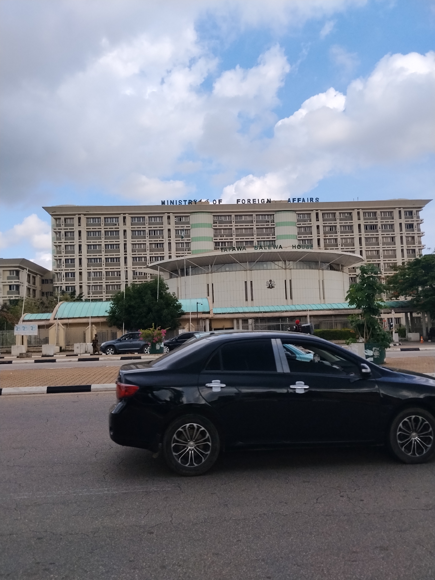 The image shows the Ministry of Foreign Affairs building in Abuja, Nigeria. The building is large and modern, with many windows. The words "MINISTRY OF FOREIGN AFFAIRS" are visible in blue letters on the top of the building. Below that, "TAFAWA BALEWA HOUSE" is also visible. The sky is cloudy with patches of blue, suggesting it is daytime. In the foreground, a black car is driving on a paved road. The road has black and white markings. There are also some trees and fences visible in the background. The overall scene appears to be a typical day in a city.