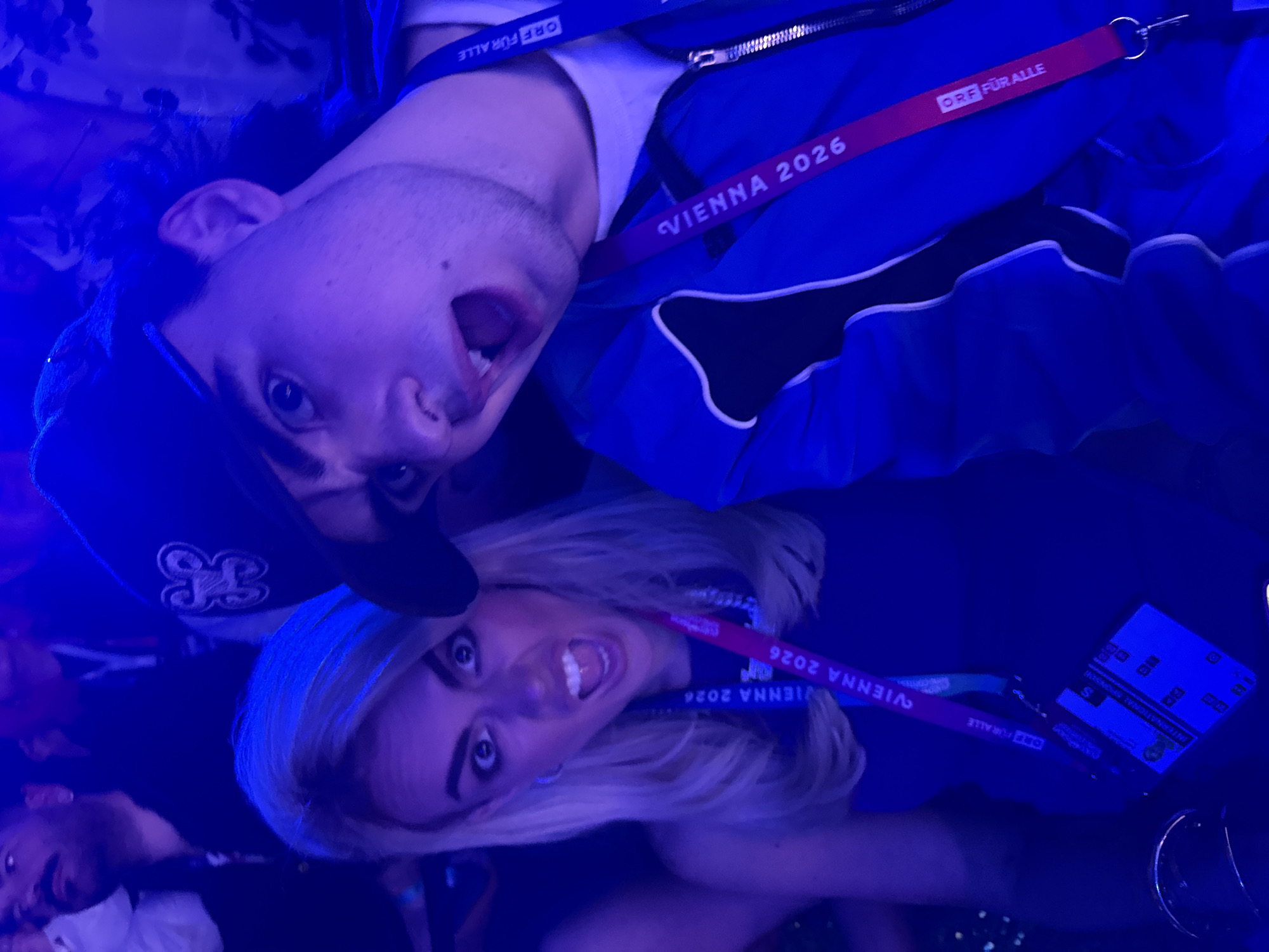 Two people are visible in this image, a man wearing a baseball cap and a blue jacket, and a woman with blonde hair. Both are wearing lanyards labeled "Vienna 2026" and "ORF FÜR ALLE." They are making exaggerated facial expressions with their mouths open. The image has a strong blue color cast. Due to the mention of "Vienna 2026" on the lanyards, it can be assumed this image was taken in Vienna, Austria, but no specific location within the city can be determined.