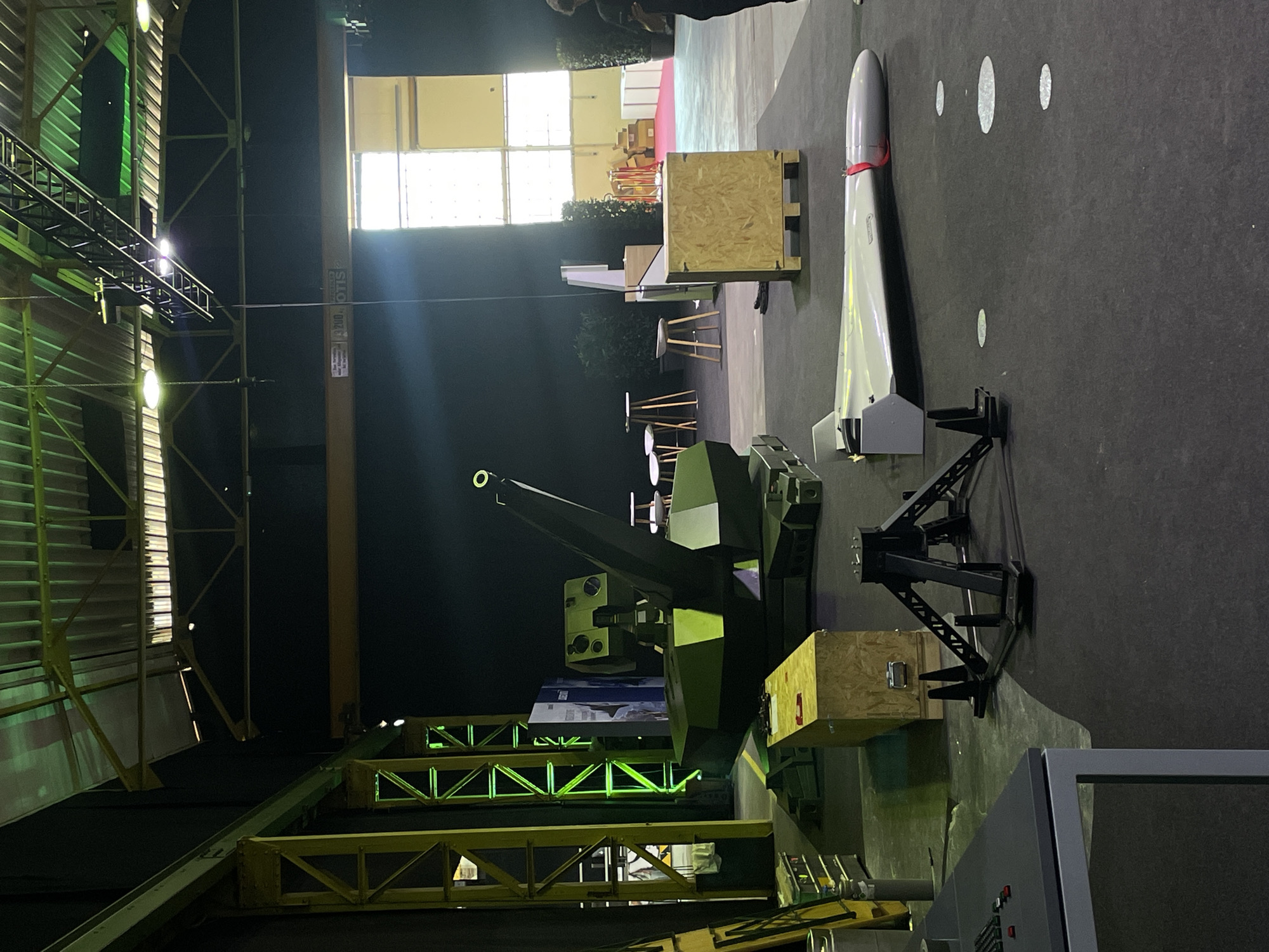 The image shows an indoor exhibition space. A green anti-aircraft gun is centrally positioned, with its barrel pointed upward. To its right is a white missile, standing vertically on a metal stand. Both items are flanked by wooden crates.
The backdrop features a well-lit window area, suggesting daylight outside. Overhead, there are industrial-looking metal beams and structures. The floor appears to be covered with a dark, possibly carpeted surface.
The environment seems to be a large hall or warehouse converted for an exhibition. The precise location is Unknown, Unknown. There are no people visible in the image.
