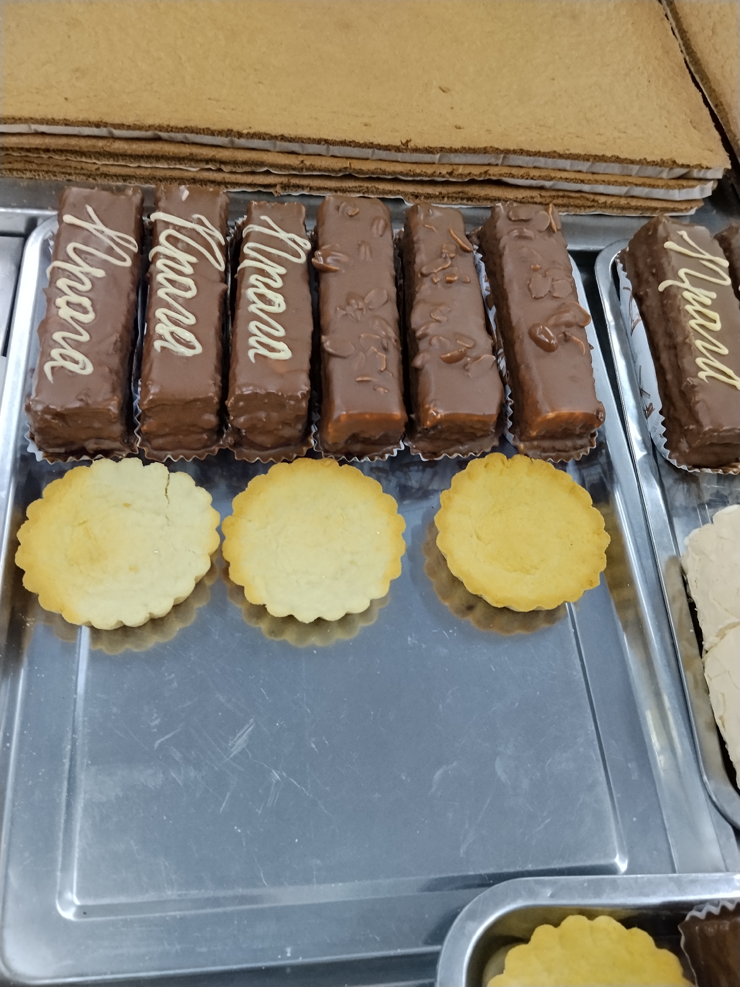 A close-up, overhead shot displays a metal tray filled with baked goods. The tray holds at least four rectangular chocolate-covered pastries, each with white lettering. Two of these pastries are mostly visible, showing the word "Lhara" written in cursive on their sides. To the left of these are two round, light-colored tarts with scalloped edges. Behind the pastries and tarts, a stack of flat, rectangular, light brown cake layers is visible. To the right of the chocolate pastries, another item is partially visible, appearing to be a white confection. The metal tray is scratched and shows signs of wear. The background is slightly out of focus. The location is Unknown, Unknown.