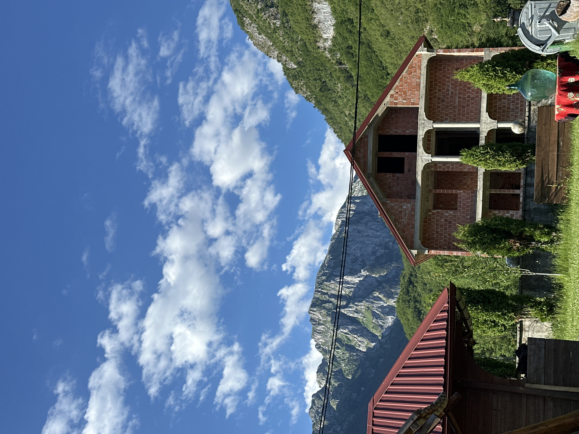 The image depicts a rural scene in Gusinje, Montenegro. In the foreground, there is a red-roofed wooden structure on the left, and an unfinished brick house with concrete columns and balconies in the center. In front of the unfinished house, a round table is covered with a red cloth, holding a large green glass bottle and a grey trash can with a lid on the right. Two conical evergreen trees stand on either side of the bottle. A wooden bench is placed behind the table. In the mid-ground, a stone wall partially separates the grassy area from the trees and vegetation. The background is dominated by lush green forested mountains under a bright blue sky with scattered white clouds. Power lines are visible across the frame. The overall impression is one of a developing rural property in a mountainous region.