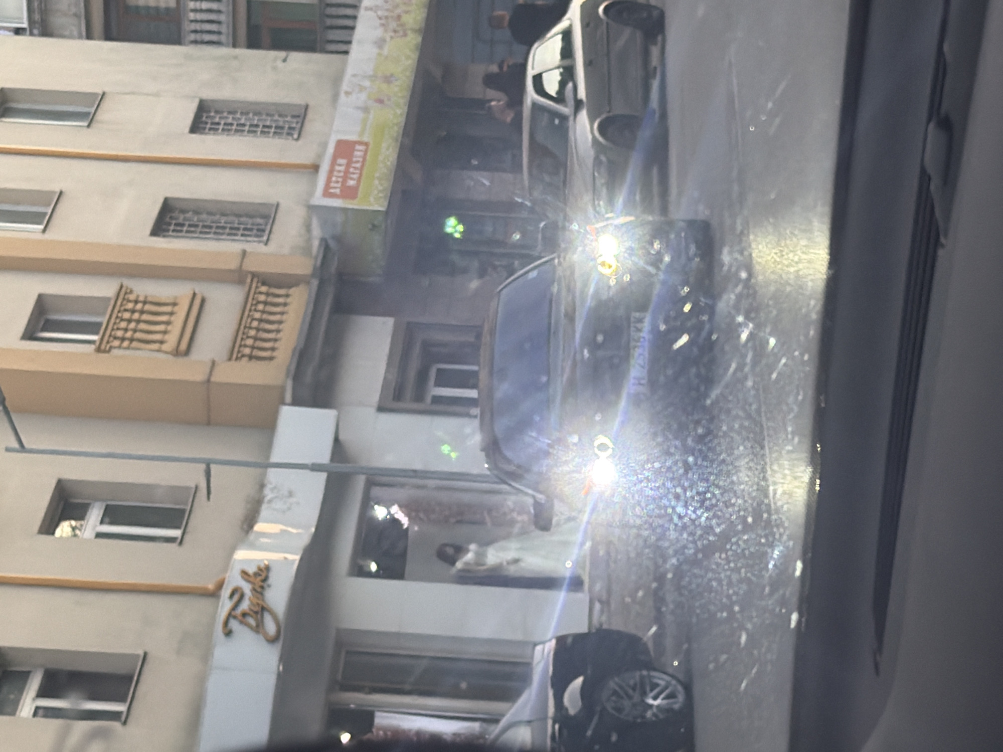 The image, captured from inside a vehicle looking through a speckled windshield, depicts an urban street scene, rotated 90 degrees clockwise. The setting is likely Shumen, Bulgaria, confirmed by the "H" prefix on a visible license plate and Cyrillic text.
In the immediate foreground, a dark-colored sedan's bright headlights create a significant glare and starburst effect on the windshield, which is covered in small droplets or specks. A partial license plate "H 7336 KK" is discernible on this vehicle. Another lighter-colored car is visible further down the street. Blurry figures of people are present on the sidewalk, suggesting pedestrian activity.
To the left, a multi-story building displays several windows, some fitted with decorative grilles or simple balconies. The ground floor of this building features a shop with a prominent, stylized golden sign in cursive script, spelling "Zayka." Further along the building, a red and white banner or sign is visible with Cyrillic text: "АПТЕКИ МАГАЗИН," which translates to "Pharmacy Shop" or "Pharmacies Stores." Inside a shop window, a figure, possibly a mannequin or person, is faintly visible.
The time of day appears to be either early morning, late afternoon, or during overcast conditions, as car headlights are active despite some ambient light. The overall scene portrays a bustling city street with a mix of traffic and pedestrian presence.