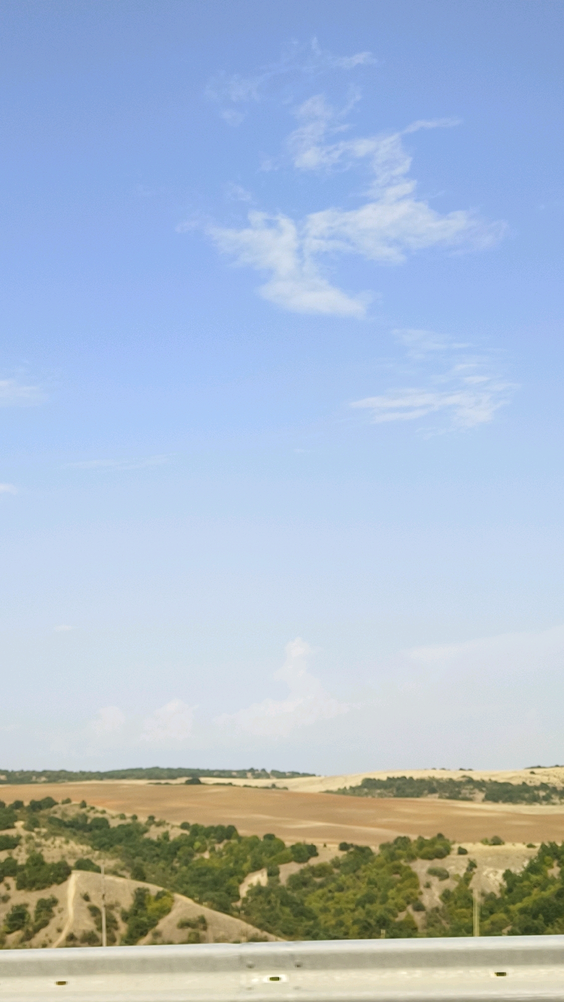 The image depicts a wide, open landscape under a vast, pale blue sky with scattered white clouds. In the foreground, a silver metal guardrail runs horizontally across the bottom of the frame. Beyond the guardrail, the terrain is varied, with rolling hills covered in dense green foliage and patches of bare earth or sand. Further in the distance, larger, flatter fields of a light brown or tan color are visible, suggesting cultivated land. The overall impression is one of a rural or natural environment. The location is specified as Novachani, North Macedonia. There are no people or discernible objects of human activity beyond the guardrail.