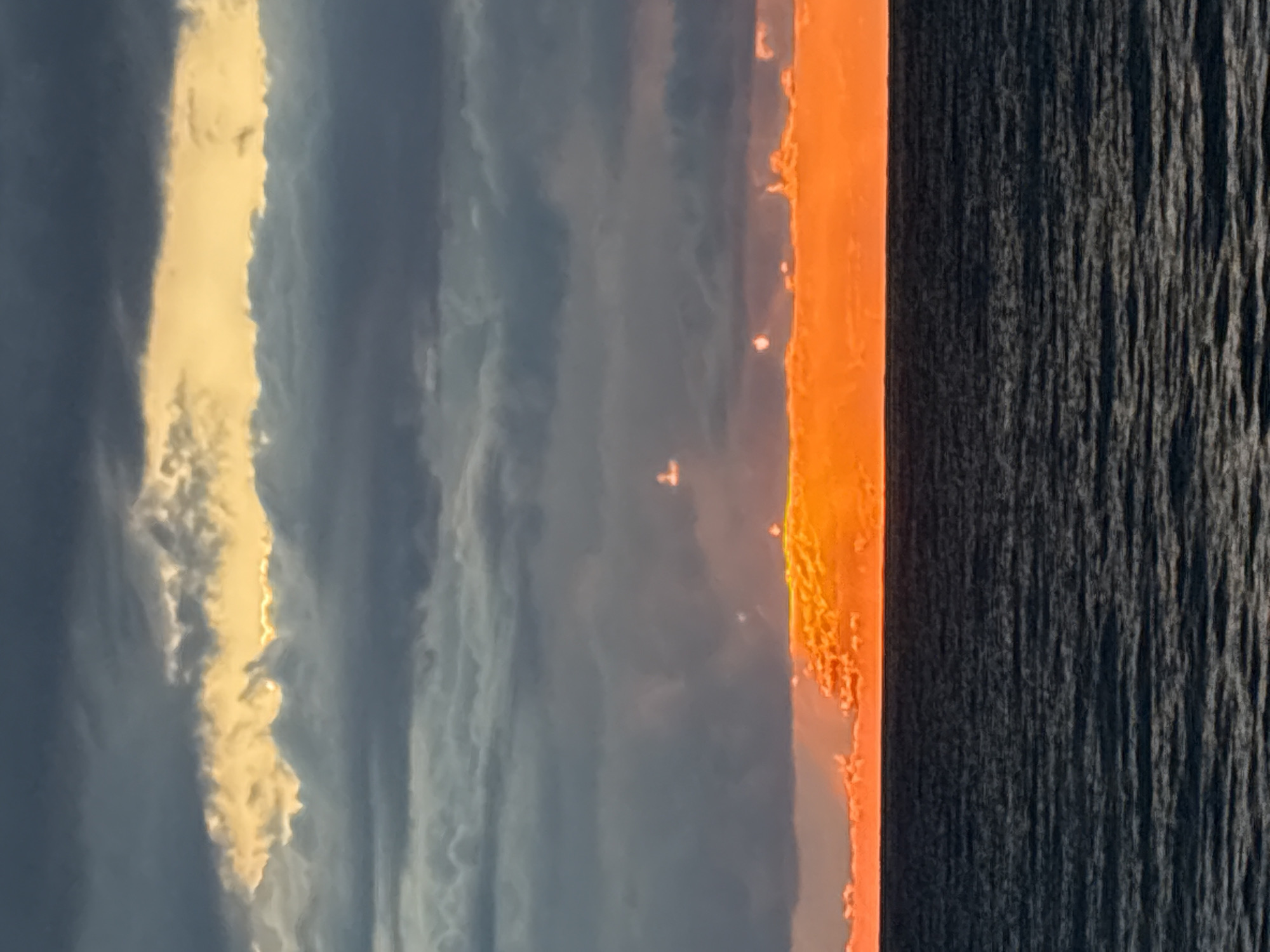 The image, taken near Bar, Montenegro, captures a dramatic sunset or sunrise over the sea. When viewed in its correct landscape orientation (rotated 90 degrees counter-clockwise), the scene is horizontally divided into three distinct elements: sky, horizon, and sea.
The upper portion of the frame is dominated by a deep blue-grey sky, indicative of either dawn or dusk. Cutting vertically (when rotated) through this darker expanse is a prominent, elongated band of bright, golden-yellow clouds, brilliantly illuminated by the low sun.
The middle section features an intensely vibrant orange-red band along the horizon, suggesting the sun is either setting or rising directly at this point. This band emits a powerful, warm glow, with small, bright specks visible along its upper edge, possibly reflections or disturbances on the water's surface near the horizon.
The lower part of the image depicts the dark, textured surface of the sea. The water displays a consistent pattern of ripples or small waves, characteristic of a body of water in low light conditions. The contrast between the brilliant orange horizon and the dark, textured sea is stark and visually striking.
There are no people or discernible objects other than the natural elements of sky, clouds, and sea. No visible text is present. The scene conveys a sense of serene natural beauty during the transition between day and night, or vice versa, over the Adriatic Sea.