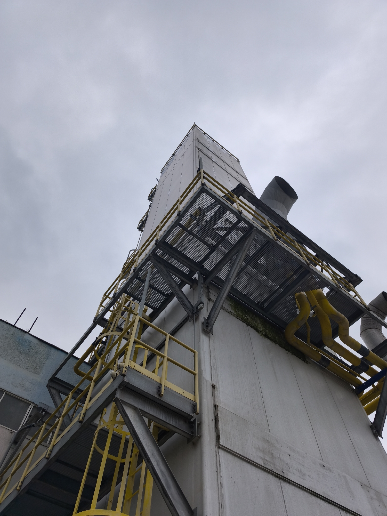 This media file presents a low-angle view of a large industrial structure under an overcast sky. The main subject is a tall, box-like building or facility constructed from light-colored, possibly white or off-white, panels.
Attached to the side of this main structure are extensive metal platforms and walkways, characterized by bright yellow railings and grated floors. These platforms are supported by robust grey metal beams and struts. Several yellow pipes and grey/silver ducts or vents are visible, some running along or exiting from the platforms. One large, curved grey duct points upwards towards the sky on the right side of the image.
A yellow caged ladder and an open metal staircase with yellow railings are prominent on the left side, providing access to different levels of the structure. A small piece of text, "L-1", is faintly visible on one of the yellow railings of the staircase.
In the background, on the far left, another building or section of a building with a blue-grey facade and some windows is partially visible, suggesting a larger industrial complex. The sky is uniformly cloudy and grey, indicating an overcast day. There are some dark, possibly green or black, stains on the white panels of the main structure, particularly below the lower platform, suggesting weathering or accumulation of grime. No human activity or specific event is taking place in the image; it is a static shot of the industrial environment.