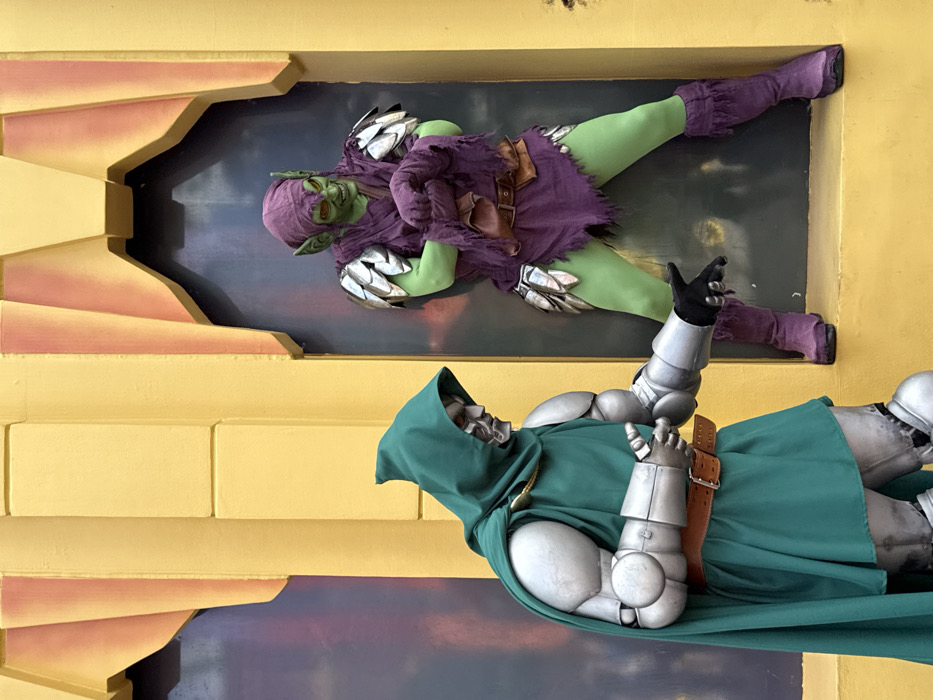 Two individuals in elaborate Marvel Comics villain costumes are depicted in an outdoor theme park setting. On the left, a person dressed as Doctor Doom stands, featuring a silver metallic mask, a green hooded cloak, silver arm and torso armor, and a brown belt. This individual extends their left hand forward with an open palm, directed towards the other subject. On the right, positioned within a dark, recessed architectural alcove, is a person costumed as the Green Goblin. This individual exhibits green skin, pointy ears, a wide grin, a tattered purple tunic, silver scale-like shoulder and thigh armor, a brown belt, and purple boots. The Green Goblin is clutching a piece of purple fabric in both hands and gazes towards Doctor Doom. The background consists of stylized yellow and orange architectural elements. This scene is located at Universal's Islands of Adventure, specifically within the Marvel Super Hero Island area, in Orlando, Florida, United States of America.