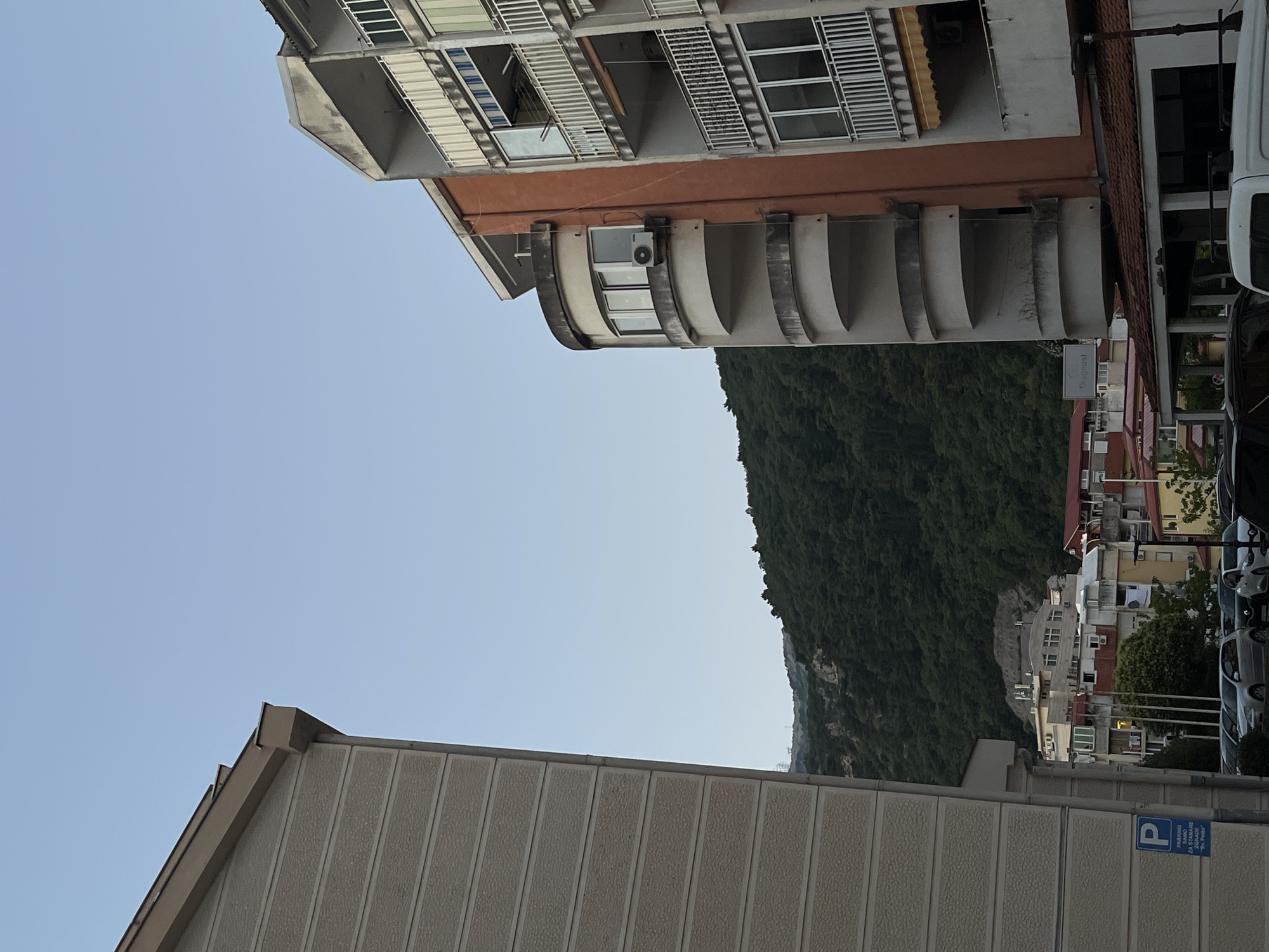 This is an outdoor, eye-level, vertical shot of a residential area in Budva, Montenegro. The sky is clear and blue, with a light haze. In the foreground, on the left, is the side of a light beige, textured building. On the right is a multi-story apartment building with an orange-tinted facade and balconies. The balconies are white with metal railings. Some of the windows have awnings. In the mid-ground, behind the apartment building, is a dense, green, tree-covered hill or mountain. Further in the distance, there are other buildings, including some with red roofs. In the lower left corner, a blue parking sign with white lettering is visible. Below the buildings, a row of parked cars can be seen. The scene appears to be set during the late afternoon or early evening. There are no people visible in the image.