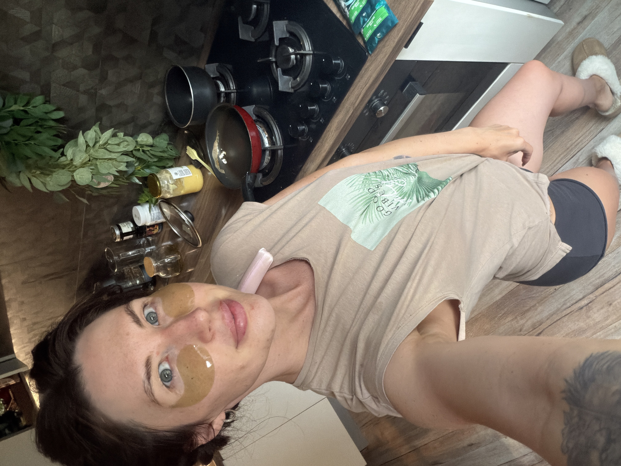 A woman with eye patches is in a kitchen, possibly in Perm, Russia, as indicated by the context. She is wearing a light brown t-shirt and dark shorts. Her hair is dark brown and pulled back. She is holding a pink object to her neck with her right hand. In the background, there is a stove with pots and pans, shelves with jars and containers, and a plant. The floor is wooden.