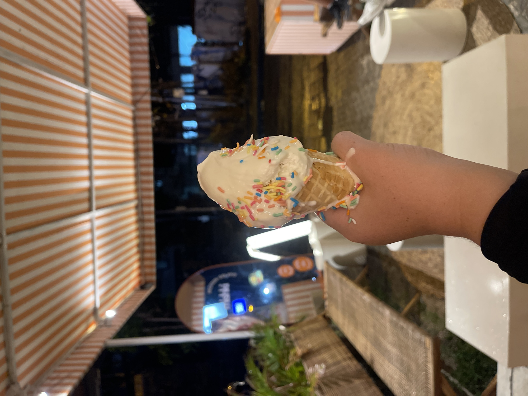 A hand holds a vanilla ice cream cone topped with colorful sprinkles. The ice cream is slightly melted, with a few drips visible on the cone. The background is out of focus but appears to be an outdoor setting at night. To the left, a building with orange and white striped awnings is visible, illuminated by string lights. In the background, there are blurry lights and structures, suggesting a street or commercial area. To the right, a white rectangular object and a roll of white paper are present. There is a sign in the background with blue and orange lettering, some of which is illegible. The overall impression is of a casual night out, possibly at an ice cream shop or street vendor.