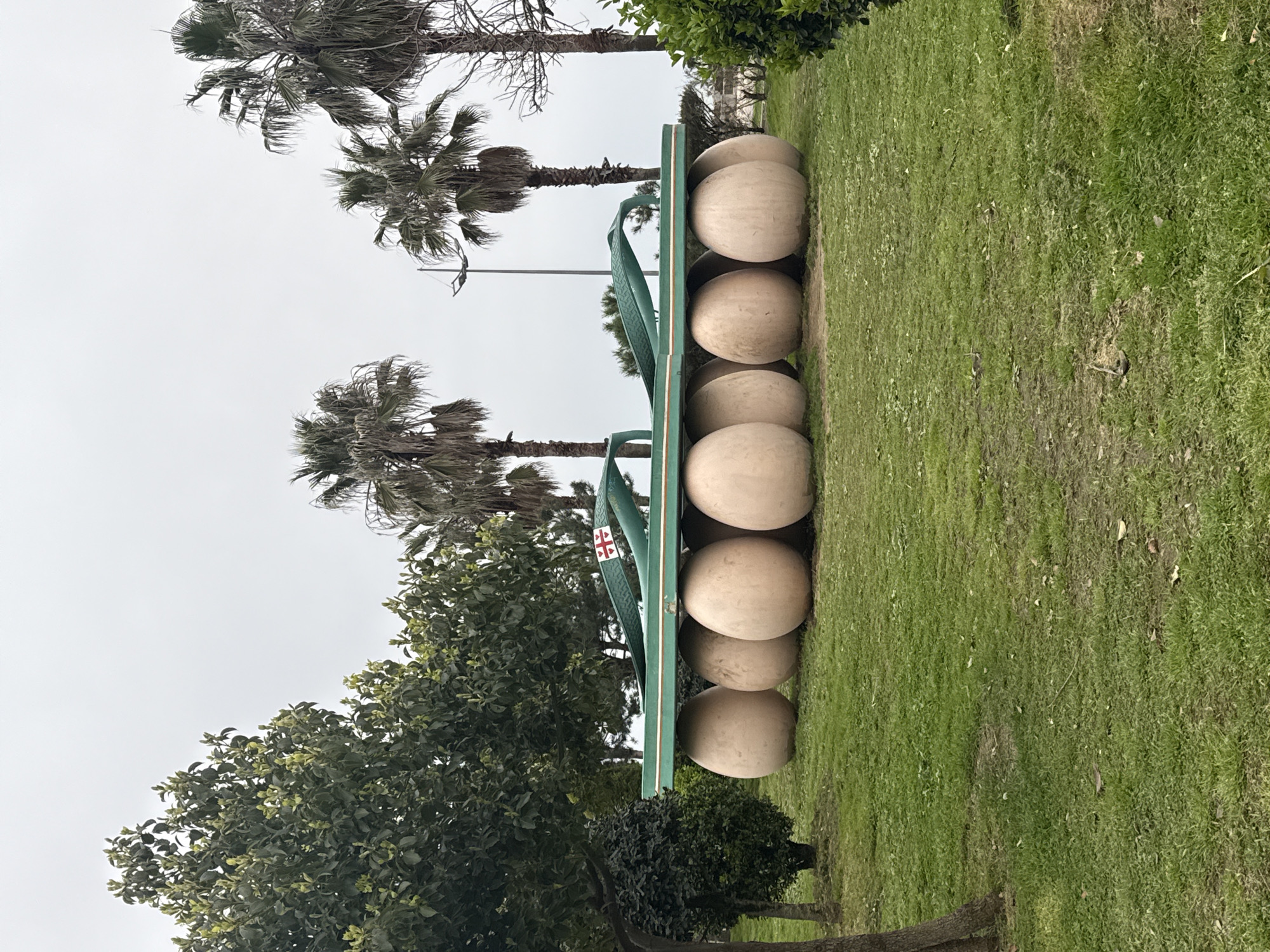 The image captures a large-scale sculpture of a flip-flop in a park setting. The sculpture is green and features a white and red symbol on the strap. The flip-flop rests on a row of large, spherical stone-like objects. Palm trees are visible in the background, as well as various other trees and green foliage. The ground is covered in grass. The sky appears overcast. The sculpture is a landmark in Batumi, Georgia.