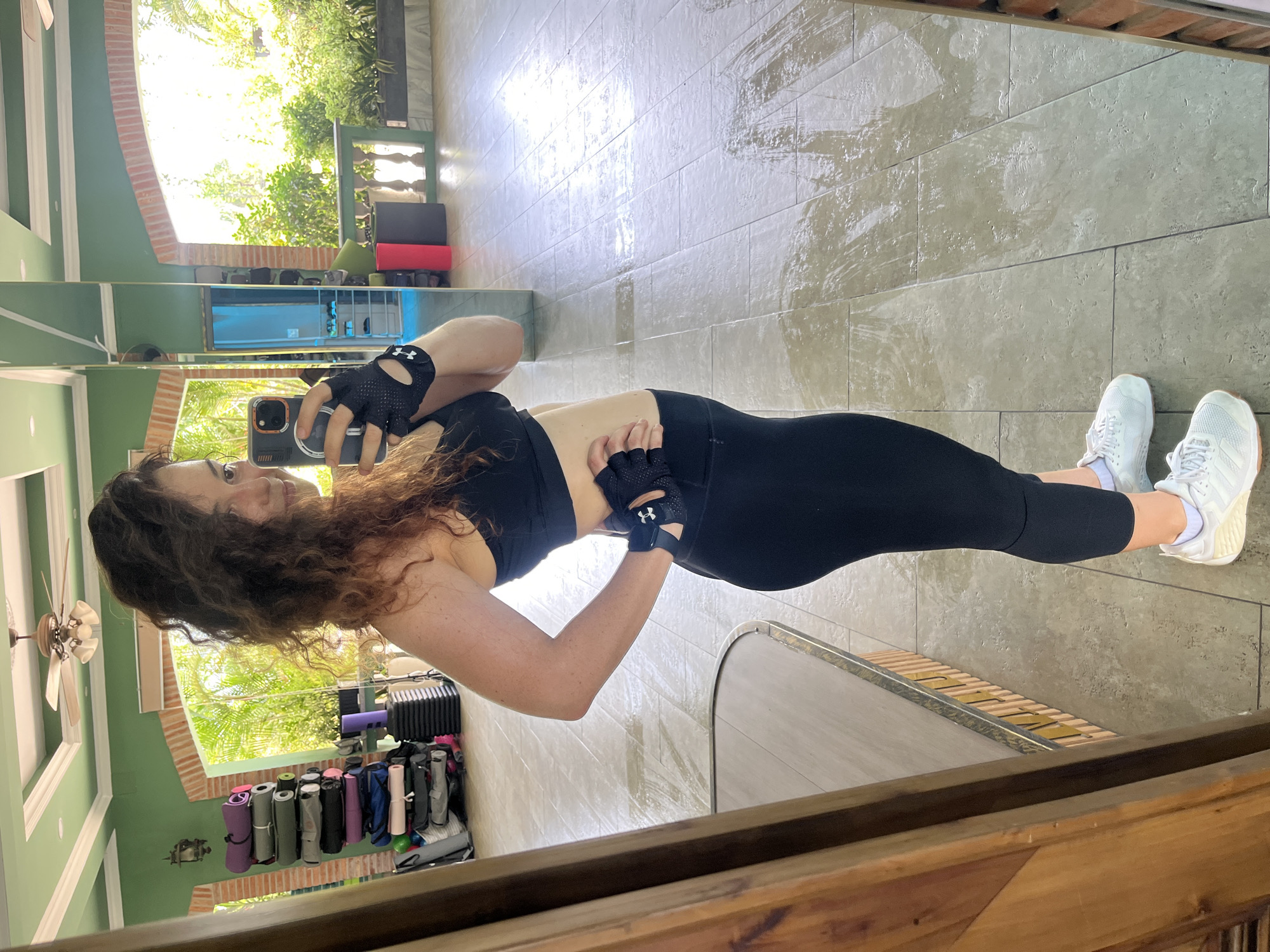 In a gym setting, a woman with curly brown hair, wearing a black sports bra and black leggings, stands in front of a mirror. She is holding her phone in her right hand, taking a selfie. Her left hand is on her hip. She is also wearing black fingerless gloves and white athletic shoes. The gym has green walls, tiled floors, and a ceiling fan. In the background, stacked yoga mats, step platforms, and outdoor greenery are visible through an open doorway or window. The floor appears wet in patches, reflecting the light. The location is Ham Thang Ward, Vietnam.
