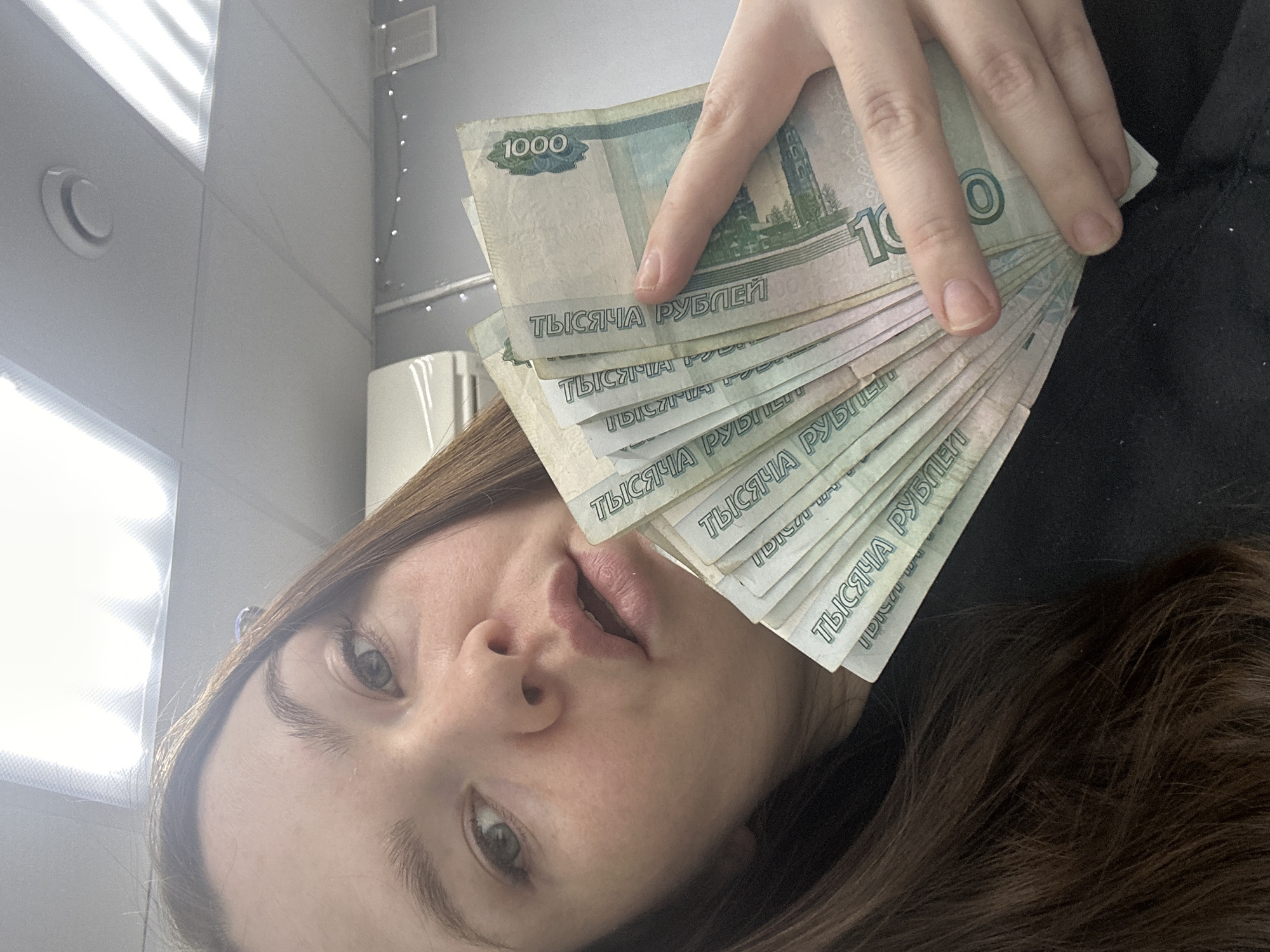 A selfie shows a fair-skinned woman holding a fan of 1000 Russian ruble banknotes. Her face is partially obscured by the money. She has light brown hair. The background shows a white paneled wall with a circular air vent, and a section of what appears to be a window with slats, suggesting an indoor setting. There is no definitive information in the image to confirm a specific location in Chaikovsky, Russia.