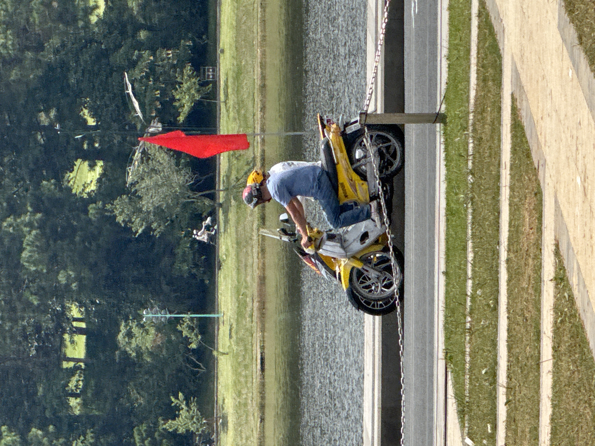 Two individuals wearing helmets are riding a yellow and white scooter along a paved road. The rider in the front is wearing a blue shirt and jeans, while the person behind them is wearing a white shirt. They are positioned on the right side of the image, with their scooter facing left. In the background, a red flag is visible on a pole. A body of water, likely a lake, is to the left of the road, with a lush green embankment and trees beyond. The overall environment suggests an outdoor setting in Da Lat, Vietnam, a city known for its scenic lakes and mountainous landscapes.