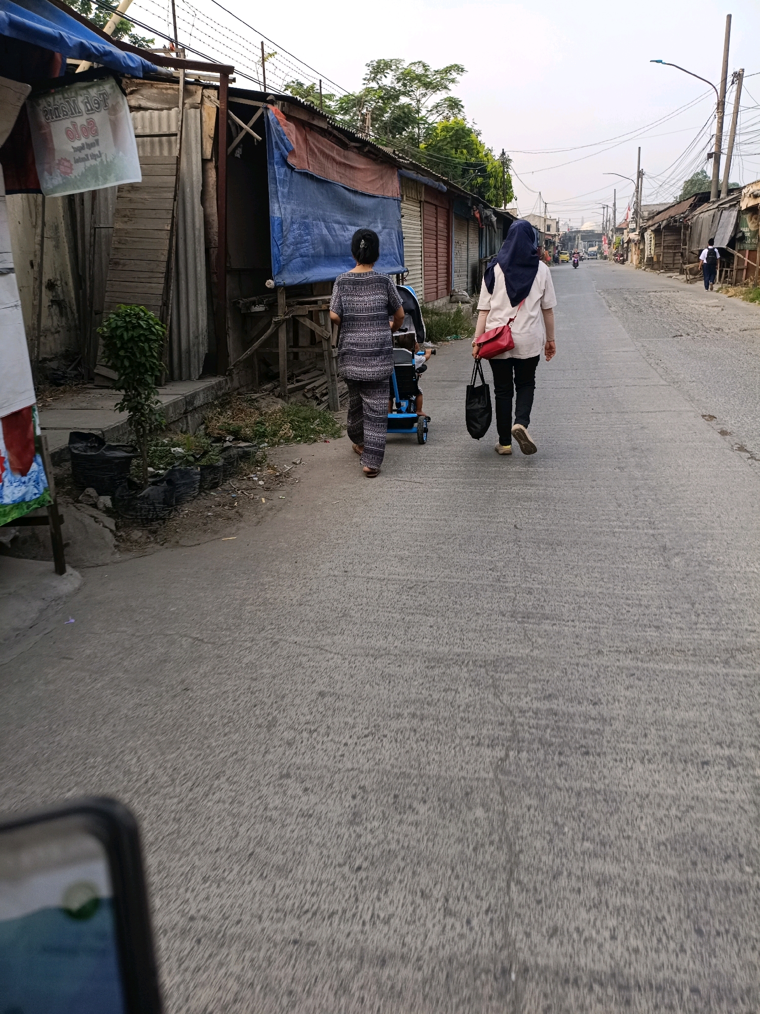 The image depicts a street scene in Tangerang, Indonesia. Two women are walking away from the camera on a paved road. The woman in the foreground on the left is wearing a patterned outfit and pushing a blue stroller. The woman on the right is wearing a light-colored shirt, black pants, and a navy blue headscarf, carrying a red purse and a black bag. They are walking past a row of makeshift shops constructed with corrugated metal and tarpaulins. In the background, the street continues with more shops on either side and power lines overhead. The overall environment appears to be an urban area with a somewhat impoverished setting.