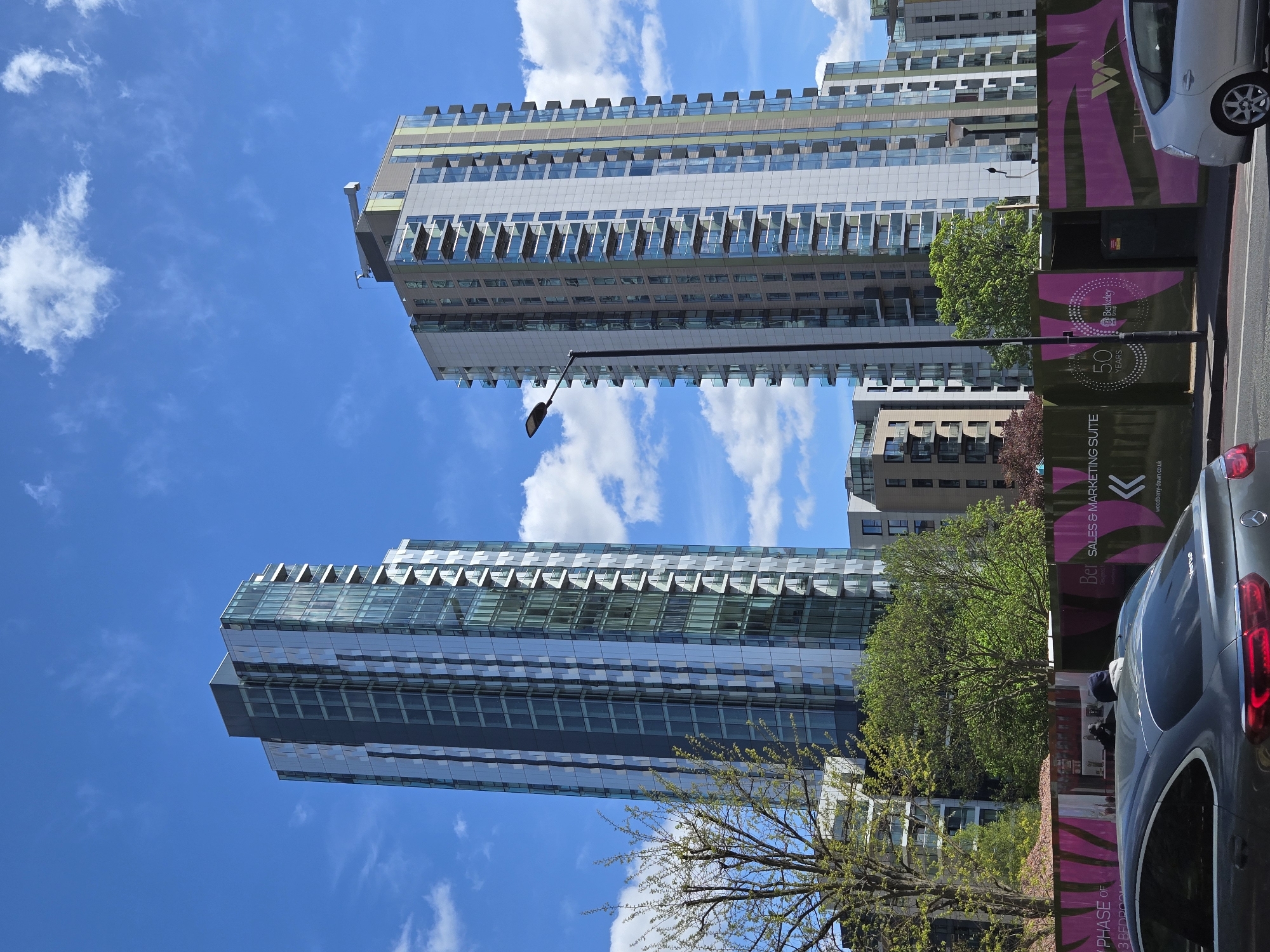 Here is a description of the image:
The image shows two tall residential buildings under a blue sky with scattered clouds. The buildings have a modern design with balconies and glass facades, reflecting the sky. In the foreground, there is a construction site with pink and green hoarding featuring branding for "Berkeley" and "Woodberry Down," indicating a sales and marketing suite. Parts of two cars are also visible along the right side of the image. The scene is an urban environment, most likely in London, United Kingdom.