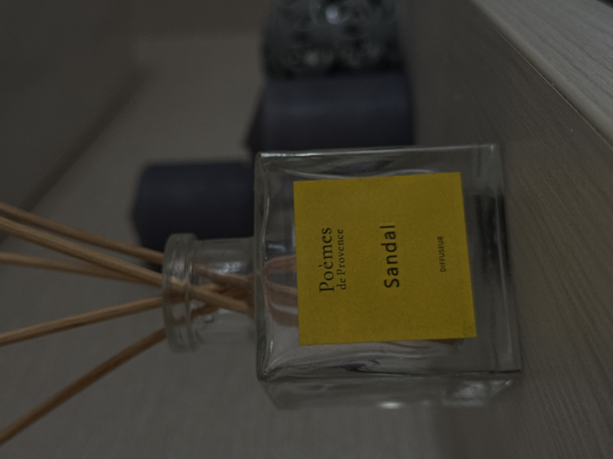 A clear glass reed diffuser bottle, featuring a prominent yellow label, is positioned centrally in a close-up, slightly overhead shot. Several light brown wooden reeds are fanned out to the left, emerging from the bottle's neck. The yellow label, oriented vertically, displays the text "Poèmes de Provence", "Sandal", and "DIFFUSEUR".
The background is softly blurred, revealing additional objects and surfaces. Behind the diffuser, out of focus, are dark grey or black items, including what appears to be a textured, spherical object and a rectangular block. The diffuser and background items rest on a light-colored surface, possibly wood or laminate, which shows a subtle grain pattern on the right side of the frame. The lighting is dim and even, characteristic of an indoor setting. No discernible activity is taking place; the image depicts a static arrangement of home fragrance items. The location context provided is Chaikovsky, Russia.