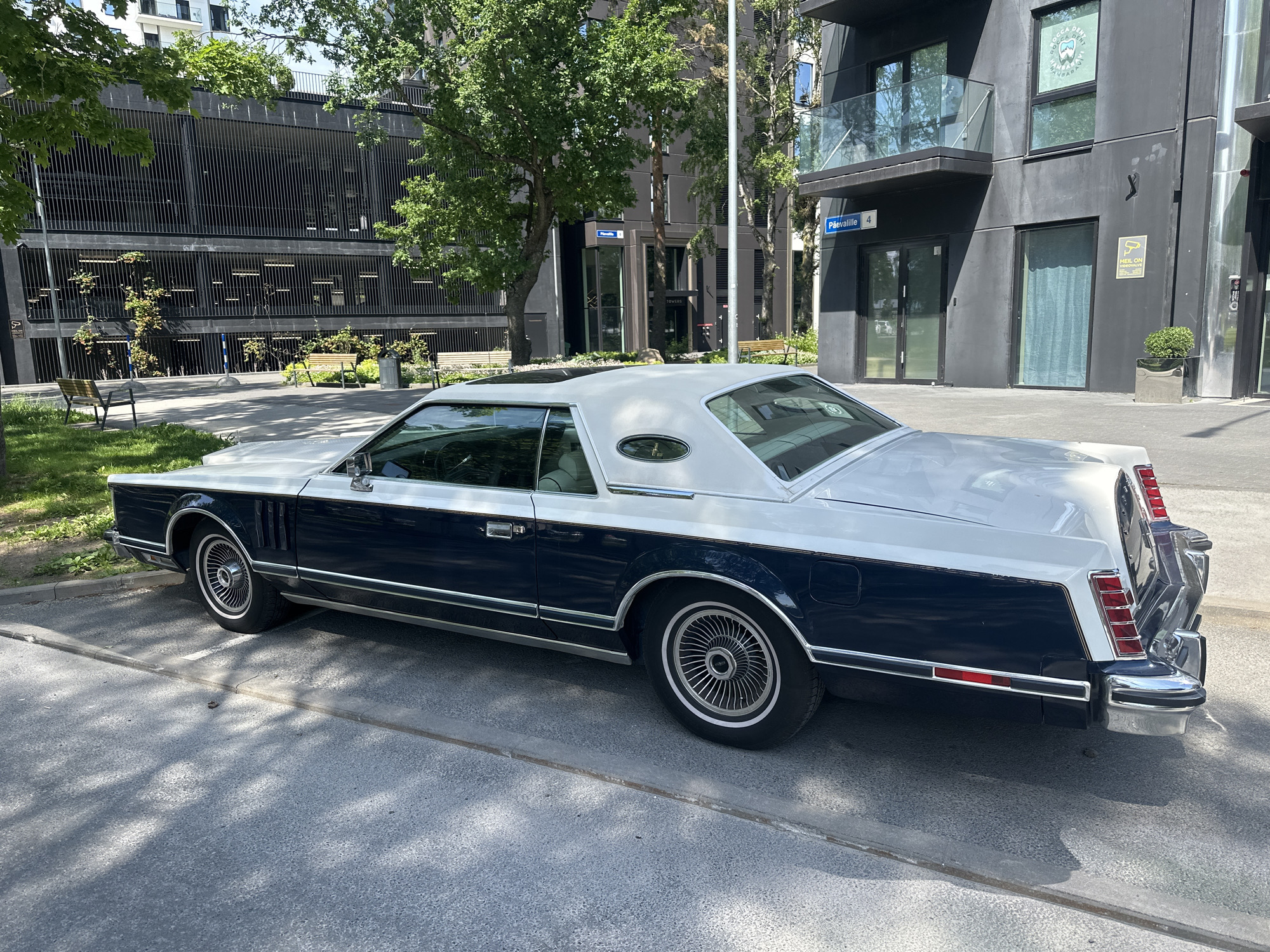 A vintage, two-tone navy blue and white Lincoln Continental Mark V is parked on a paved surface in front of a modern, dark-colored building. The car is viewed from its rear-right quarter. The building features multiple stories with balconies and large windows. In the background, a multi-level parking garage with a slatted facade is visible, as are trees and street signs. One sign reads "PÄEVALILLE 4". The scene appears to be a street or public space in Tallinn, Estonia, under a clear, sunny sky.