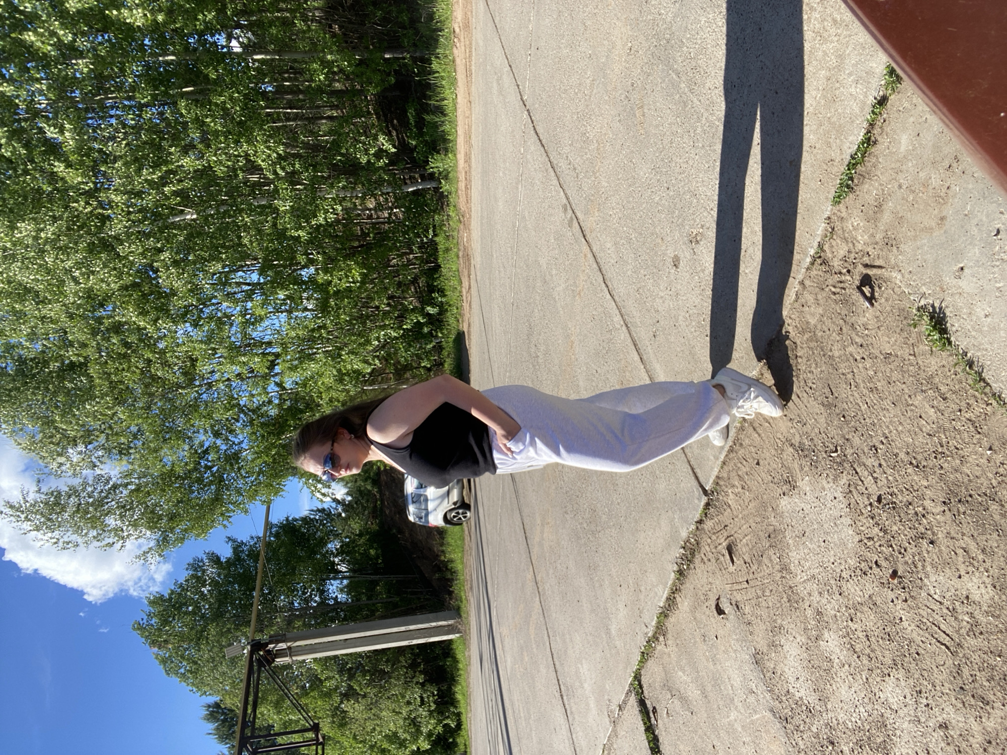 A young woman stands on a cracked concrete surface, likely a road or sidewalk, with her left hand in her pocket. She is wearing sunglasses, a black tank top, light-colored sweatpants, and white sneakers. Her shadow extends along the pavement to the right.
In the background, there is a dense forest with a blue sky visible above. A metal structure, possibly a utility pole or a small tower, can be seen amid the trees. A white car is parked on the road behind the woman. The setting appears to be a rural or suburban area bordering a forest. The location is identified as Chaikovsky, Russia.