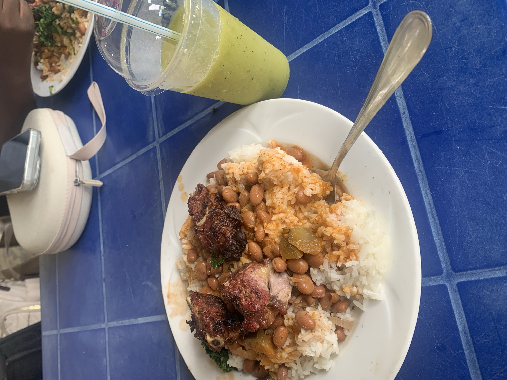 The image shows a table with two plates of food and a drink. On one plate is a meal of rice, beans, and what appears to be roasted pork, accompanied by a spoon. Another plate contains a mixed rice dish with vegetables, and next to it is a plastic cup with a green smoothie or juice and a straw. There is a small white bag with a light pink strap on the table. The table surface is blue with a tiled pattern. This scene is likely set in Dar es-Salaam, Tanzania, as per the prompt's request. No people are fully visible, only a small portion of a person's arm and hand are on the table near the plate with mixed rice and vegetables.