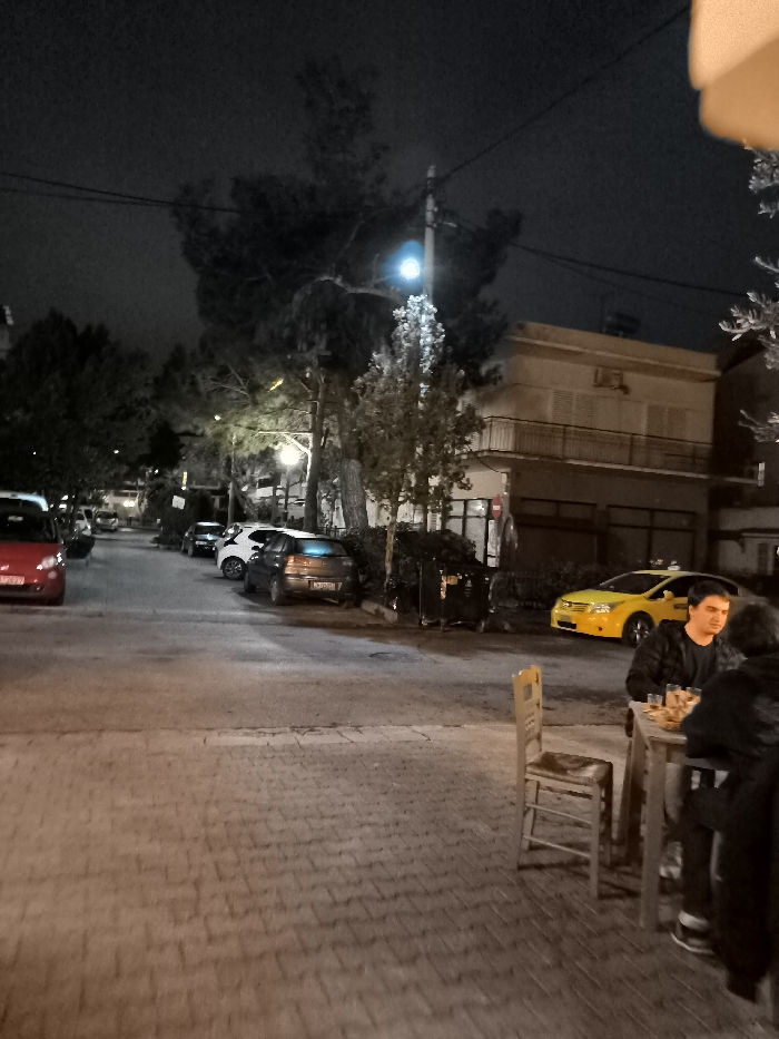 This nighttime image depicts a quiet street scene in Nea Erithrea, Greece. In the foreground on the right, two people are seated at an outdoor wooden table with chairs. One man, wearing a plaid shirt, is clearly visible facing left, while the second person has their back to the viewer. Small plates of food and drinks are on their table, indicating a relaxed social setting, possibly at a cafe or restaurant.
The street, paved with interlocking bricks, extends into the background where several vehicles are parked. A red hatchback is visible on the far left, followed by a white and a dark gray hatchback further down the road. A bright yellow taxi is prominently parked on the right side of the street, near black trash bins. Tall trees with varying foliage line the street, interspersed with power poles and lines. Streetlights cast a blue-white glow in the mid-ground and warmer light further back, illuminating the scene. Buildings with balconies are visible behind the trees and cars, contributing to the urban environment. The overall atmosphere is tranquil. No readable text is discernible in the image.
