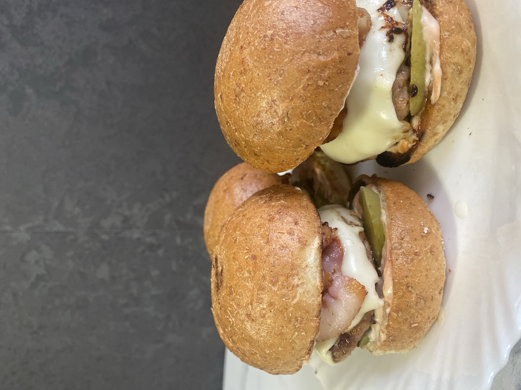 Two small sandwiches sit on a white plate. The sandwiches appear to be made with whole wheat buns and contain cheese, meat, and pickles. The image has a gray background. There is no information to infer that this image was taken in the city of Izhevsk, Russia.