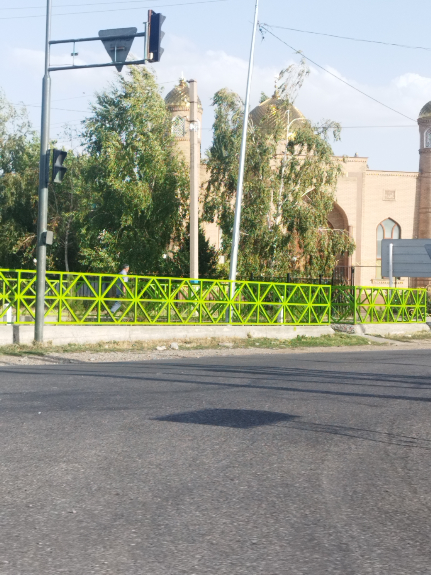 The image shows a street scene in Зертас ауылдық округі, Kazakhstan. In the foreground, there is an asphalt road with tire tracks and shadows cast from an unseen object. Behind the road, a bright lime green metal fence with a geometric pattern runs horizontally across the frame. A person wearing a blue shirt and grey pants is walking behind the fence, facing to the left. Beyond the fence, a row of trees with green foliage is visible. In the background, a large building with a golden dome and brick walls stands under a clear blue sky with some clouds. Traffic lights are mounted on a pole on the left side of the image.