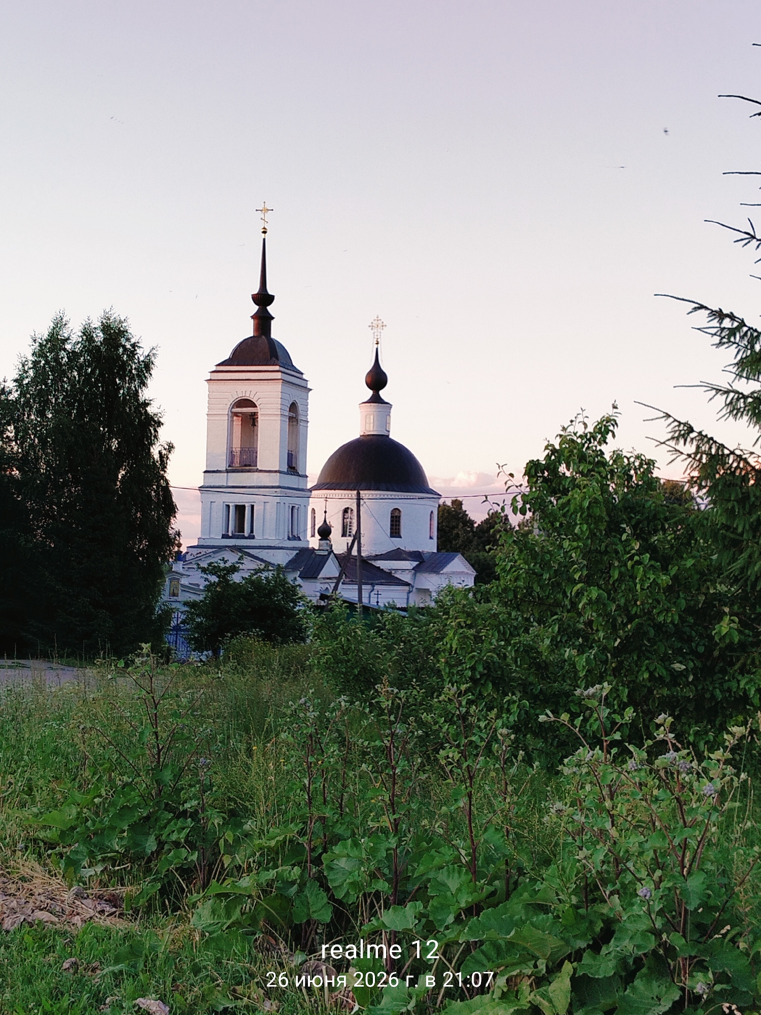 The image shows a view of the Church of the Assumption of the Blessed Virgin Mary in Novoe, Russia, as captured on June 26, 2026, at 21:07. The church, a white structure with a dark dome and a bell tower, is partially obscured by lush green trees and foliage in the foreground and to the sides. The sky is a pale, clear hue, suggesting either dawn or dusk. The scene is dominated by natural elements, with the architecture of the church serving as the central subject. The camera's metadata indicates it was captured with a Realme 12 device.
