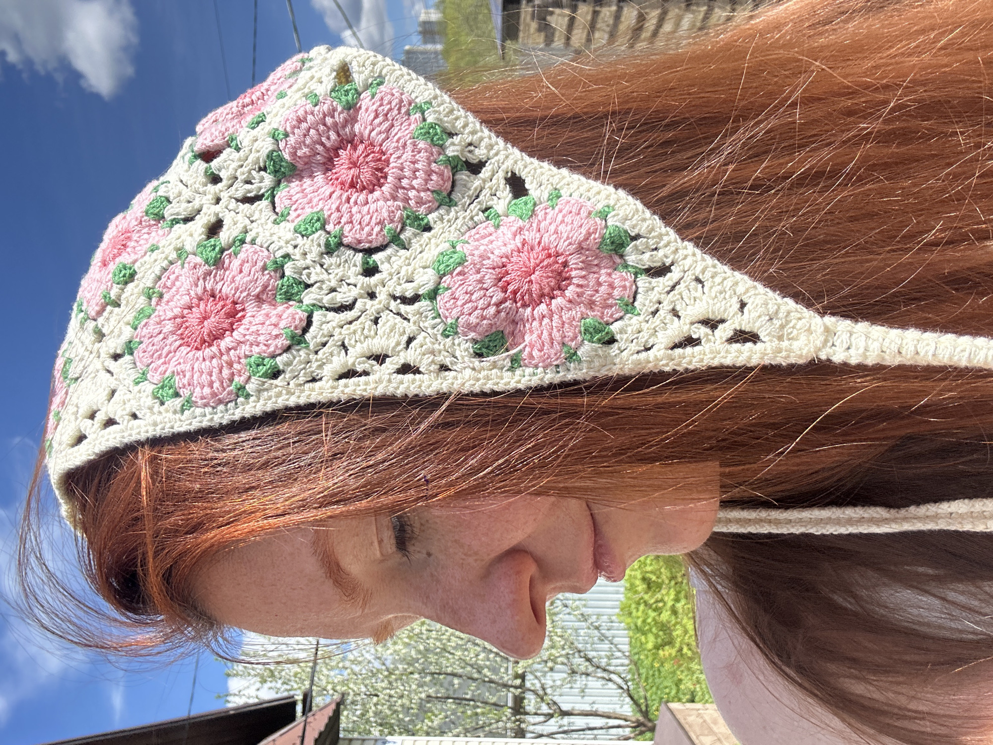 A side-view close-up photograph features a red-haired woman wearing a crocheted head covering with pink floral designs. The covering is primarily off-white with pink flowers and green accents. The woman's face is in profile, showing her forehead, eye, nose, and lips. Her skin has visible freckles. The background includes a blue sky with clouds, suggesting an outdoor setting. Faint outlines of buildings are visible in the distance above the horizon. The scene does not provide information to identify the location as the city of Nizhny Novgorod, Russia.