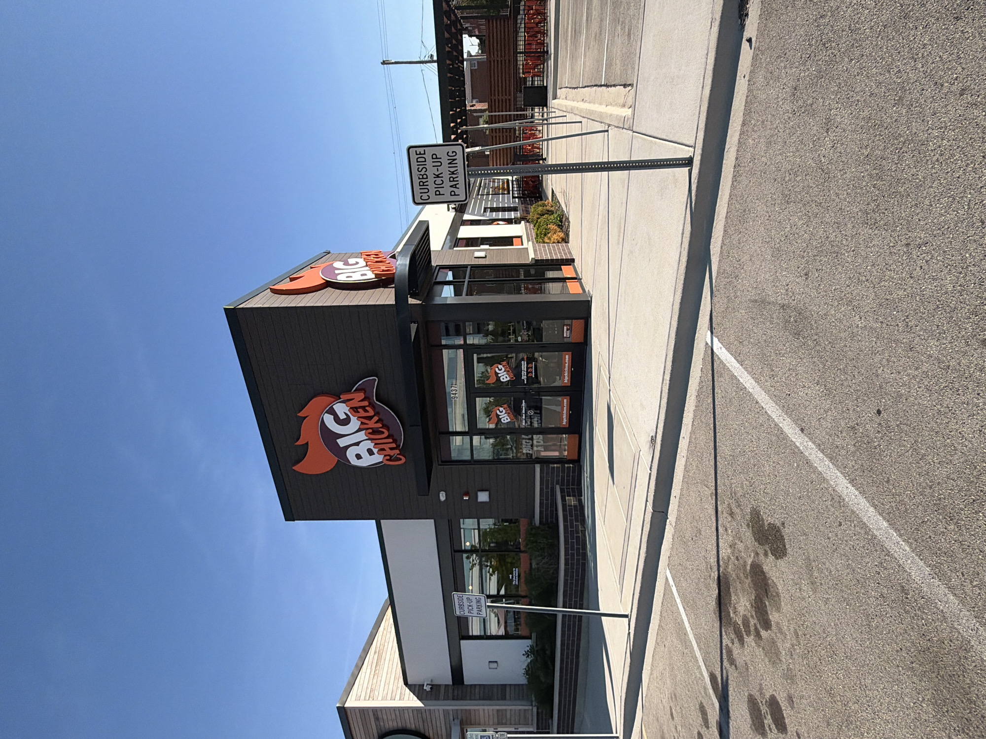 The image depicts a Big Chicken restaurant located in Rosemont, Illinois, United States of America. The building has dark gray siding and a prominent Big Chicken logo with an orange chicken head. The restaurant has glass doors and windows with signage. A "Curbside Pick-Up Parking" sign is visible near the entrance. The ground surface is asphalt with painted parking lines and some oil stains. The sky is clear and blue.