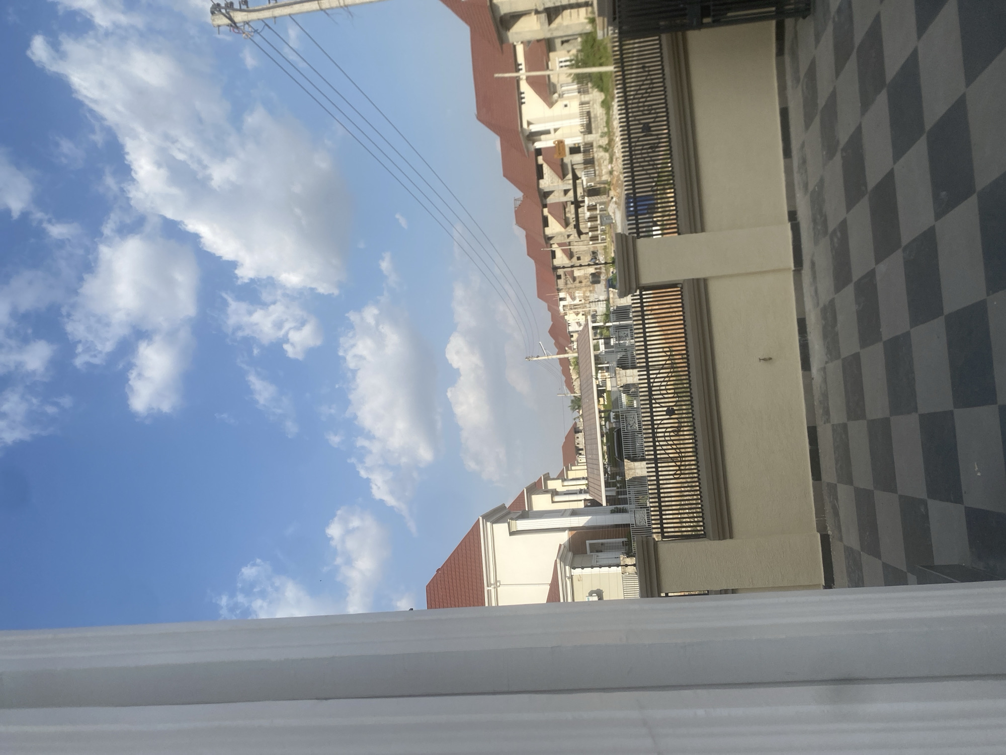 The image, taken from a slightly elevated viewpoint, shows a street scene in Iddo Tudunwada, Nigeria, characterized by newly constructed, uniform residential houses with red-tiled roofs. The sky occupies a significant portion of the frame, revealing a blue expanse dotted with scattered white clouds. Power lines stretch diagonally across the sky. A tiled walkway with a black and white checkerboard pattern is visible in the foreground, along with a portion of a light-colored exterior wall and a wrought-iron fence. The view looks down the street showing a row of similar houses. The lower part of the image is blurry, showing the frame of what seems to be a window. There are no people visible in the image.