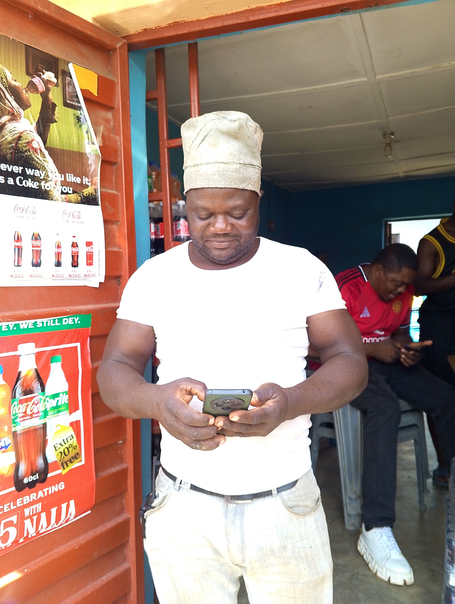 A man wearing a white t-shirt and light-colored pants stands in front of a small shop, looking down at his phone. He wears a light-colored cap. Behind him, other men are visible, one in a red Manchester United jersey and another in a black and yellow basketball jersey. The shop appears to sell beverages, with posters and bottles of Coca-Cola and Sprite visible. The environment seems to be outdoors or a covered outdoor space, with wooden and blue structures. The lighting suggests daytime.