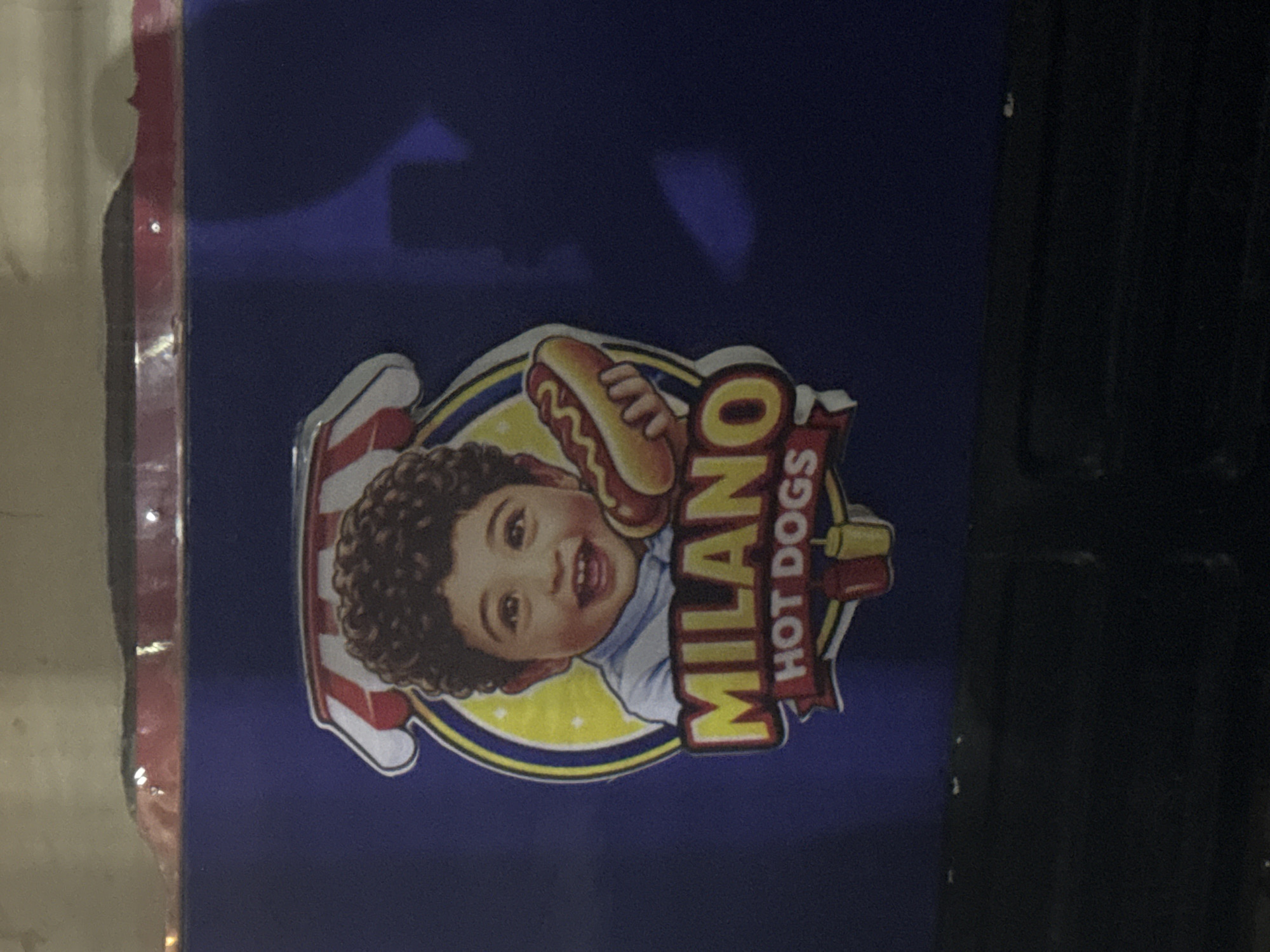 The media file features a cheerful product logo set against a dark blue background. The central element of the logo is a smiling young child, likely a boy, with curly brown hair, looking directly at the viewer. The child's left hand (partially visible) holds a hot dog in a bun, positioned near their head. Behind the child, there's a bright yellow arc and a design incorporating red and white stripes, possibly resembling a flag or a decorative border. The child is depicted wearing a light blue t-shirt.
To the right of the child, the brand name "MILANO" is prominently displayed in large yellow letters, with "HOT DOGS" written in smaller red letters vertically below it. The overall design suggests a food product advertisement, conveying happiness and appeal through the child's joyful expression. The dark blue background is uniform. On the left edge of the image, a lighter, possibly beige or white vertical surface is visible, while the right edge shows a dark, vertically ribbed or paneled structure, hinting that the logo is affixed to a larger object like a display case or a refrigerator. There are no direct visual cues within the image to specify its location in Abuja, Nigeria, but this is the provided context.